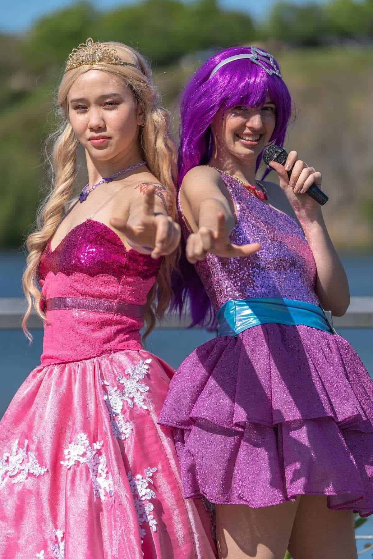 Two people in pink and purple costumes pose outdoors with one holding a microphone, both extending an open hand toward the camera.