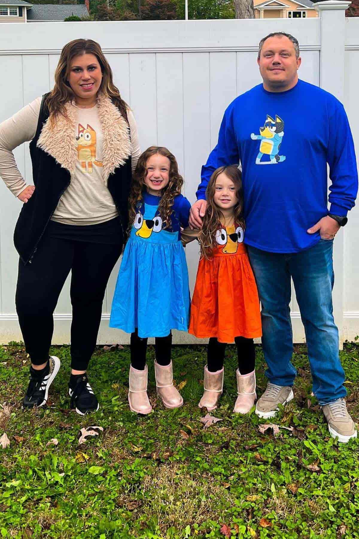 A family of four stands outside in matching Bluey-themed outfits, with the parents in shirts and the two children in blue and orange dresses, smiling at the camera.