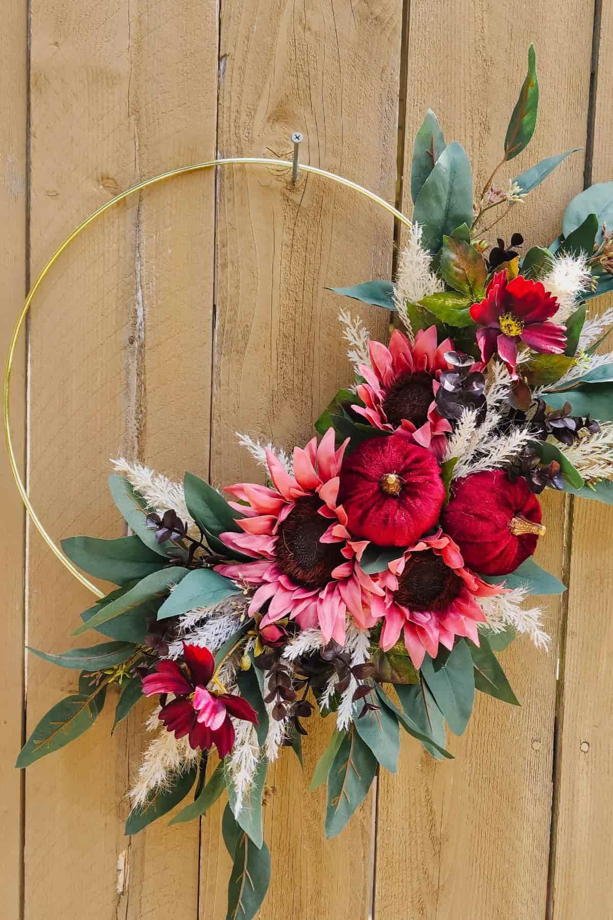 A gold hoop wreath decorated with artificial red flowers, pink sunflowers, green leaves, and white dried grasses hangs on a wooden fence.