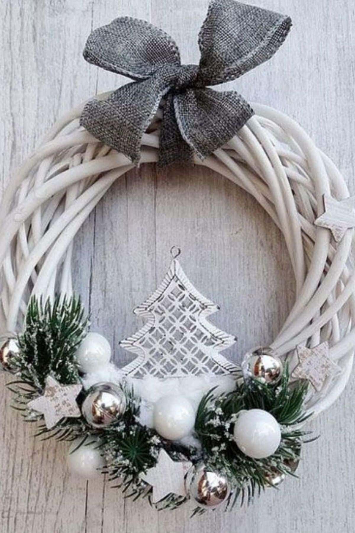 White wreath with pine branches, white and silver ornaments, star decorations, a metal tree centerpiece, and a gray fabric bow on a light wood background.