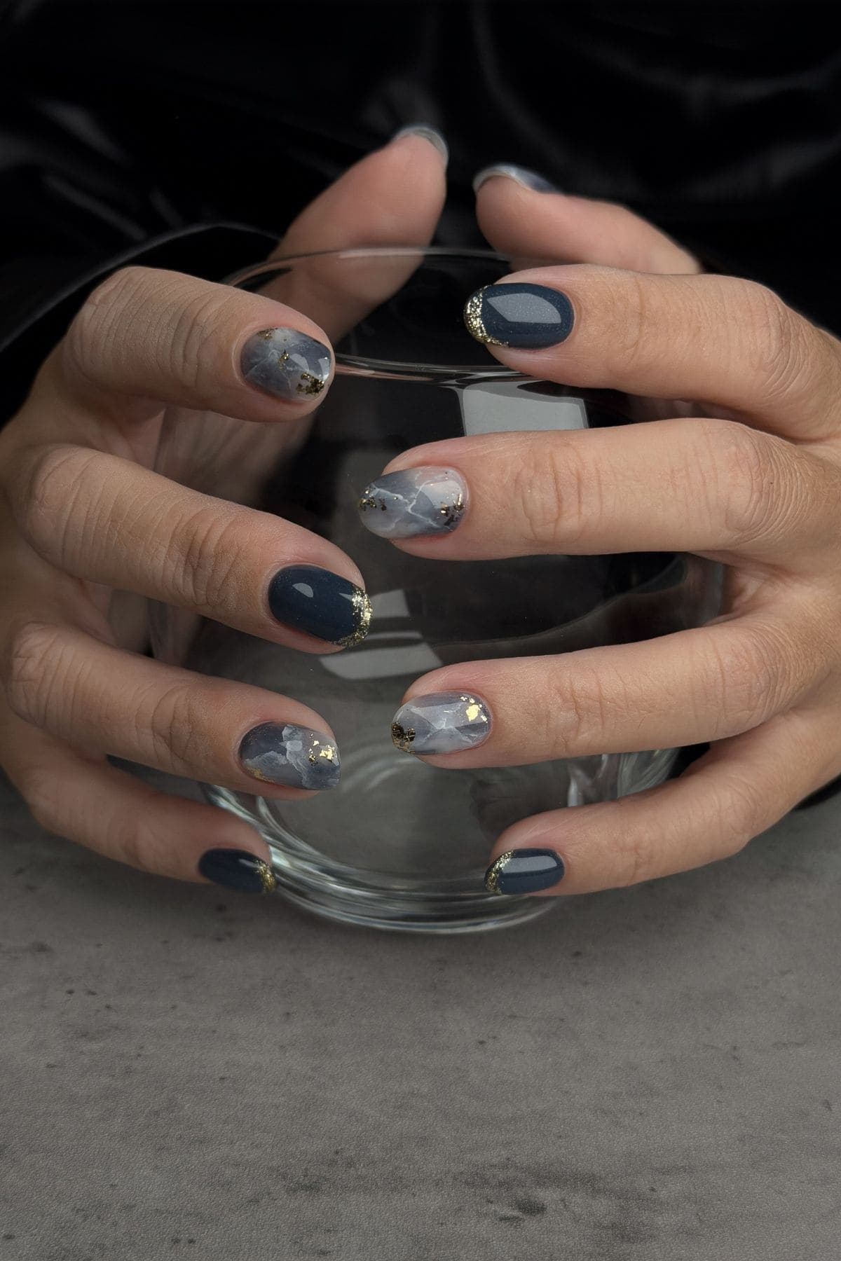 Hands with manicured nails featuring dark blue polish, marble design, and gold accents, holding a clear glass bowl on a gray surface.
