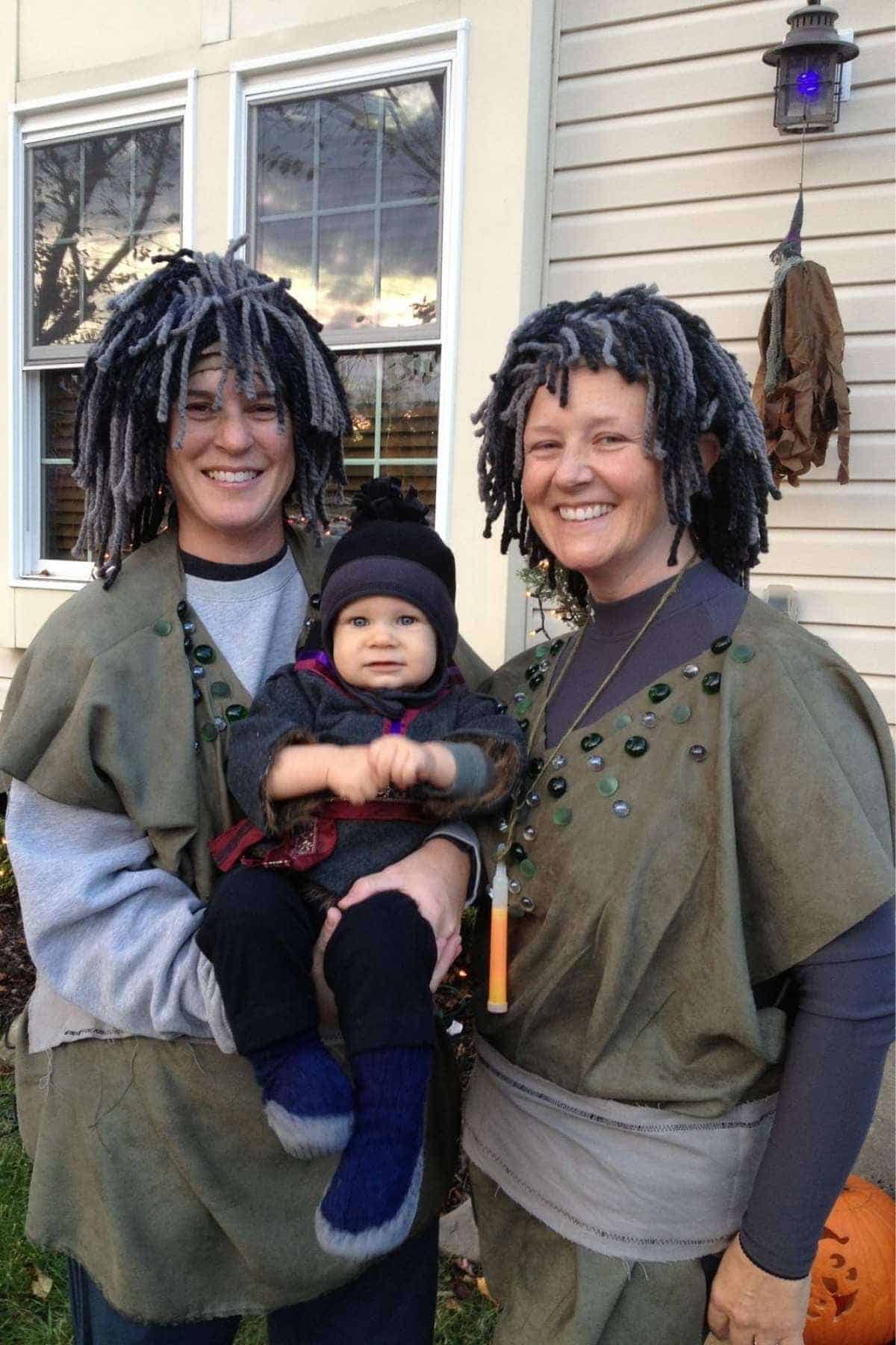 Two smiling adults in matching costumes with yarn wigs hold a bundled-up baby outside a house. The adults wear green outfits with buttons and accessories.