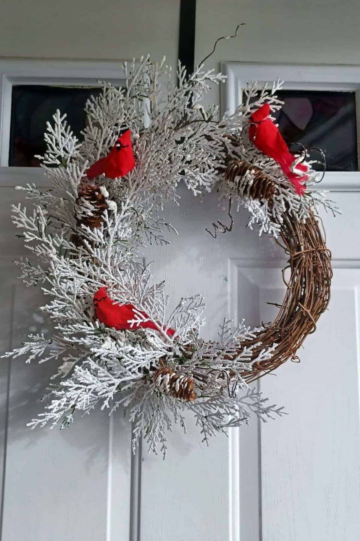 A winter wreath with frosted branches, pinecones, and three artificial red cardinals is hanging on a white door.
