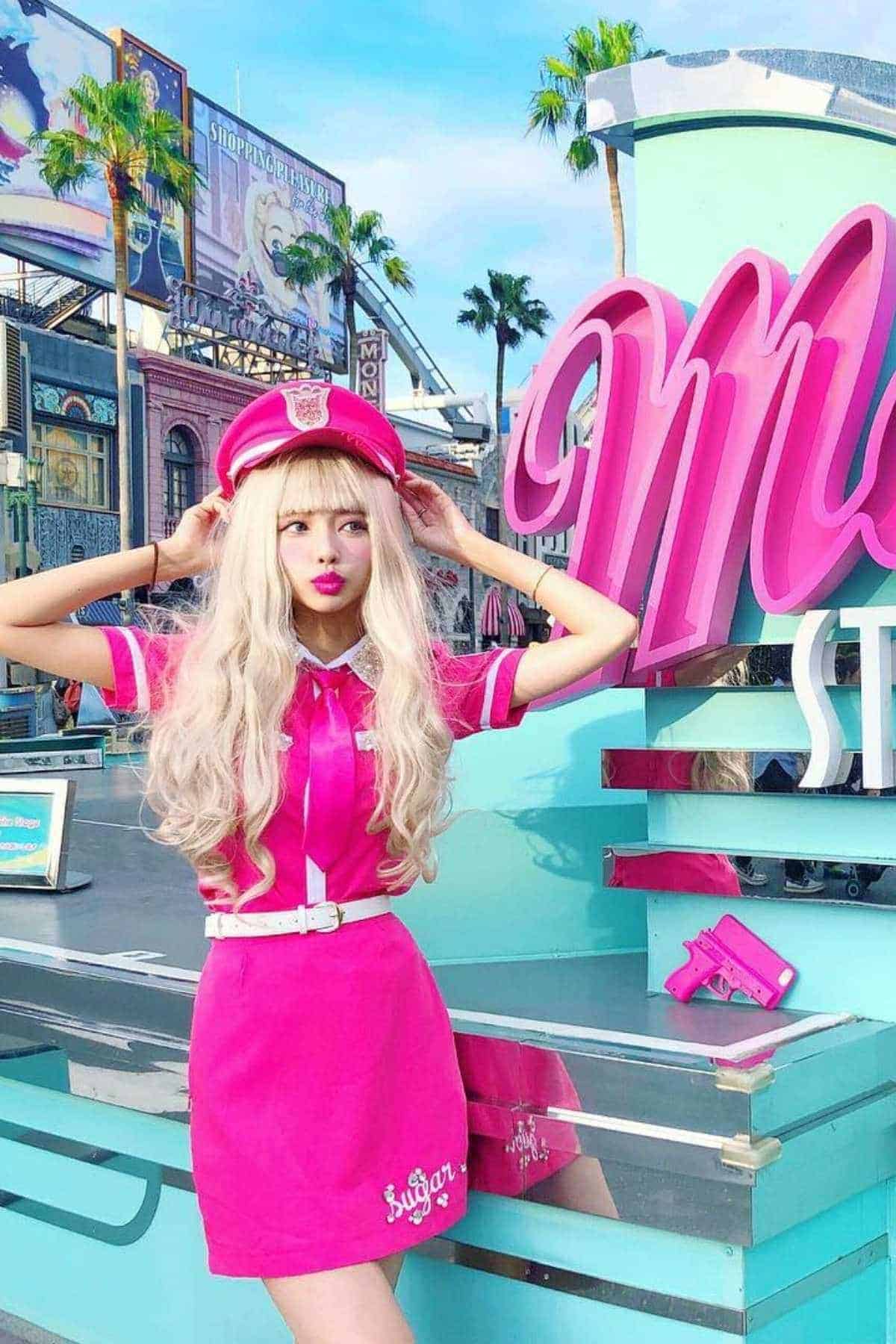 Person with long blonde hair in a bright pink outfit and hat poses in front of a pastel-colored sign and city backdrop with palm trees and billboards.