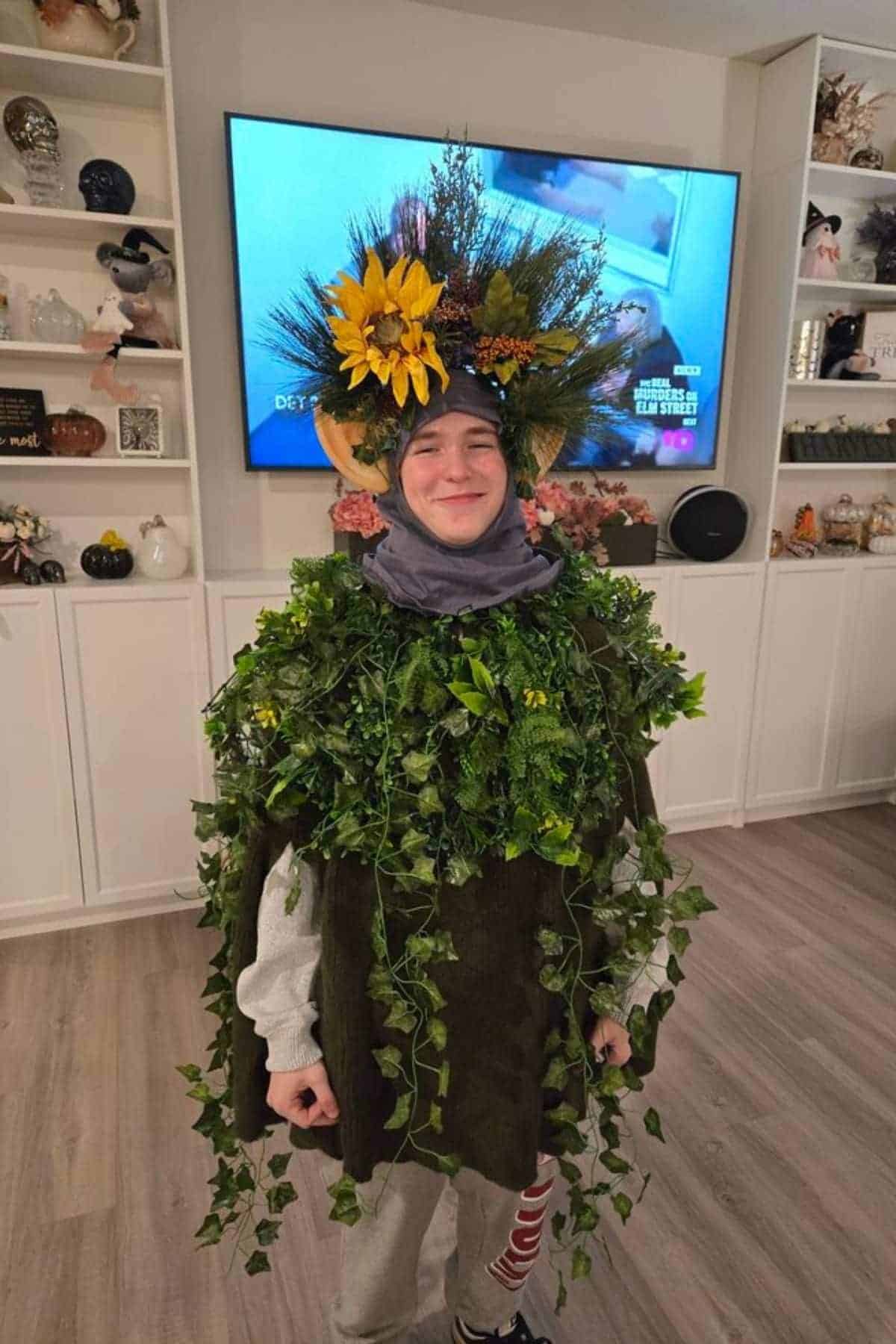 Person wearing a costume covered in faux greenery, leaves, and sunflowers, standing indoors with shelves and a TV in the background.