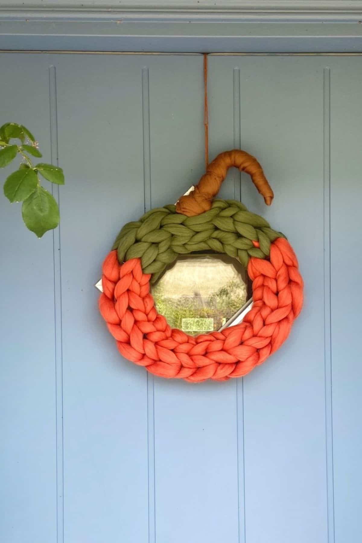 A round mirror framed with chunky, braided orange and green yarn to resemble a pumpkin, hangs on a light blue door.
