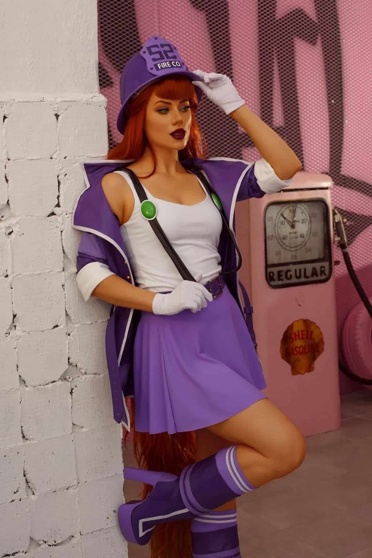 Person in a purple and white costume with suspenders and a helmet leans against a white brick wall; vintage gas pump in the background.