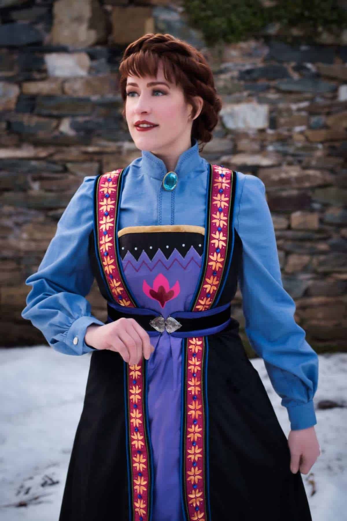 A woman wearing a blue and purple folk-style dress with decorative patterns stands outside in front of a stone wall and snow.
