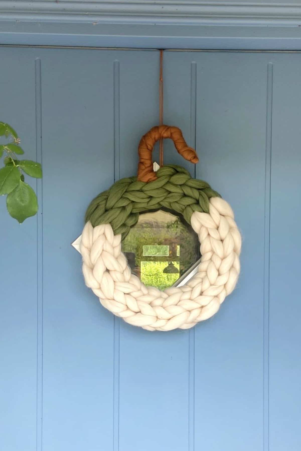 A chunky knit wreath with green, cream, and brown yarn shaped like a pumpkin hangs on a light blue door.