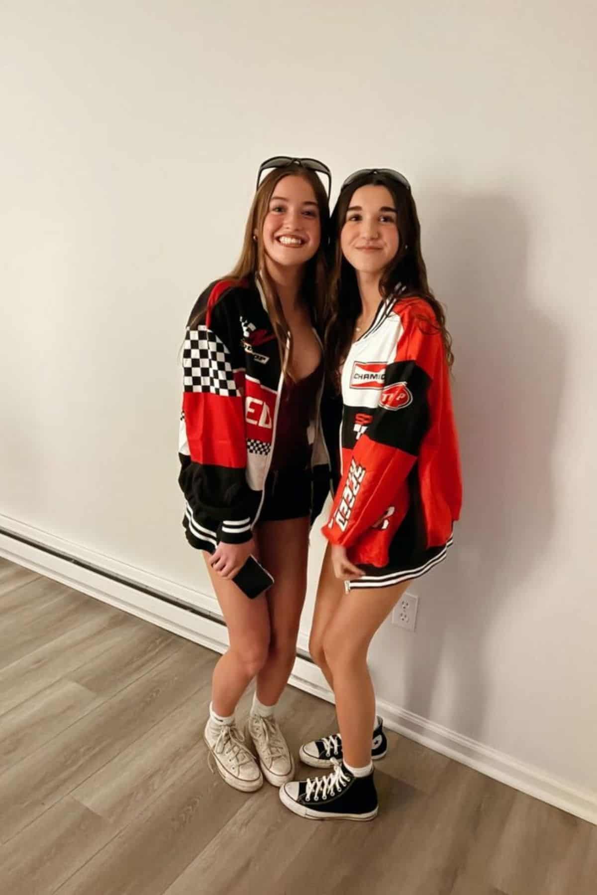 Two young women stand side by side smiling, wearing racing jackets, shorts, and sneakers, in a room with a plain wall and wood flooring.