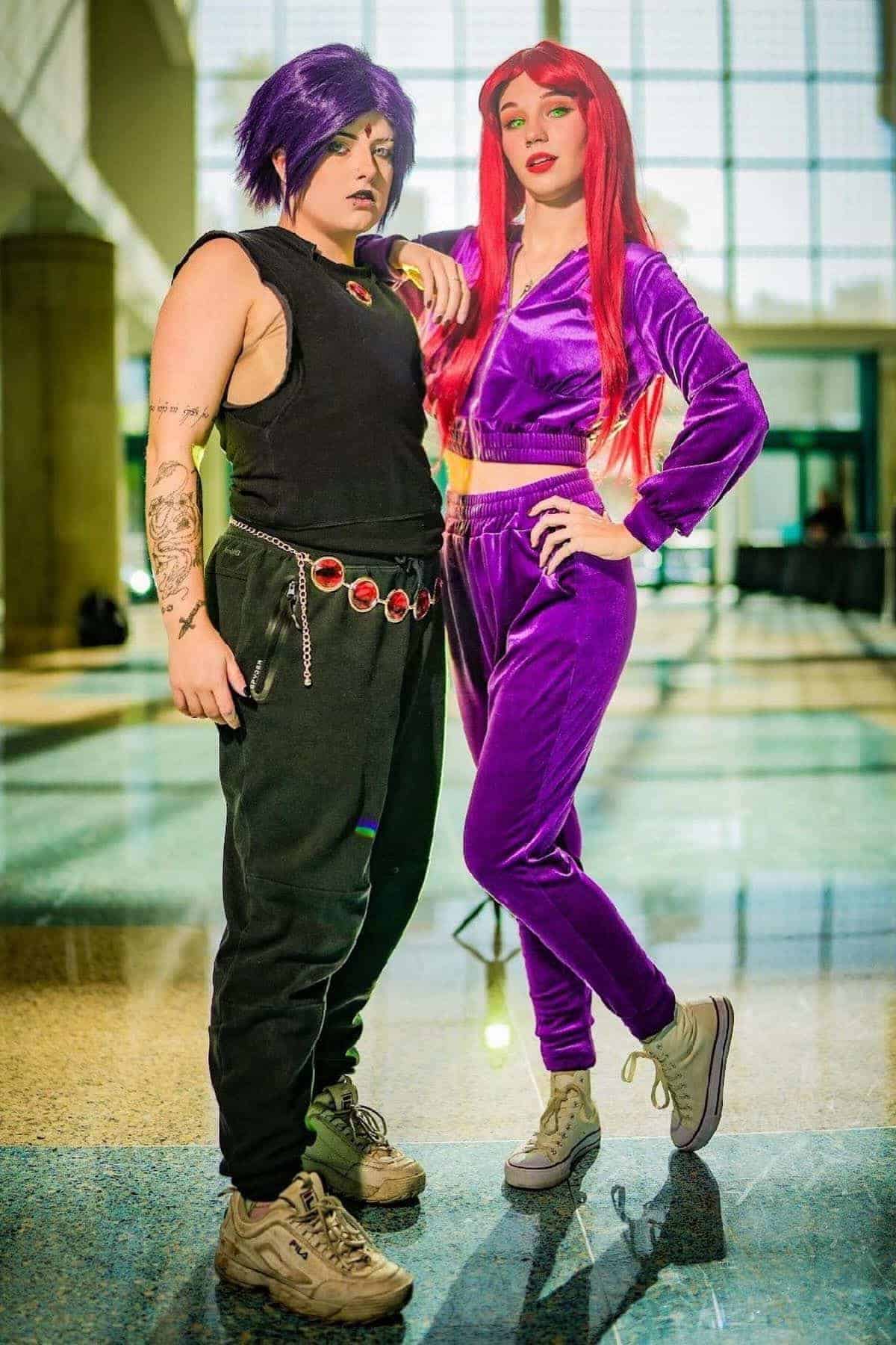 Two people in cosplay stand indoors, one in black with purple hair and red belt, the other in a purple tracksuit with long red hair. Both wear light-colored sneakers.
