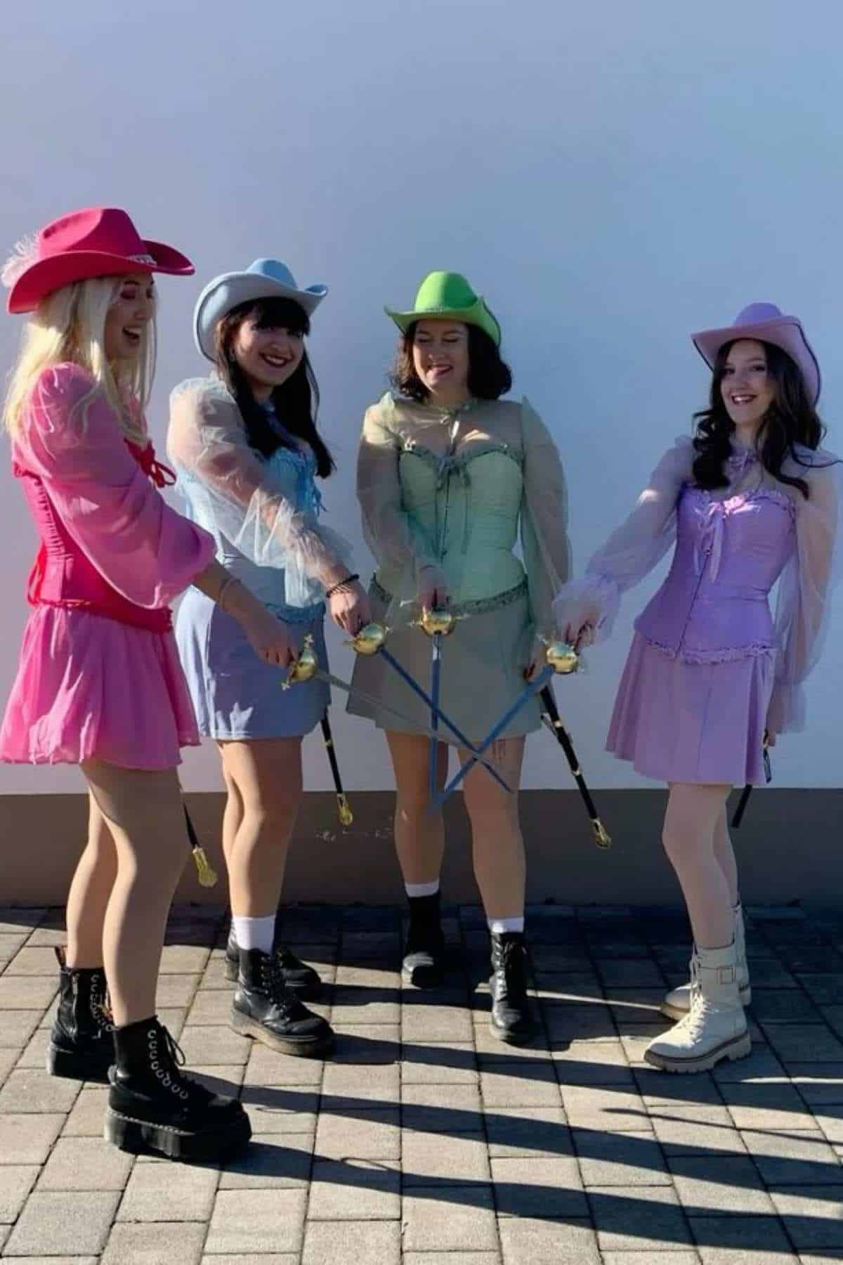 Four women in colorful dresses and cowboy hats stand in a circle outdoors, each holding a sword toward the center, smiling and posing together.