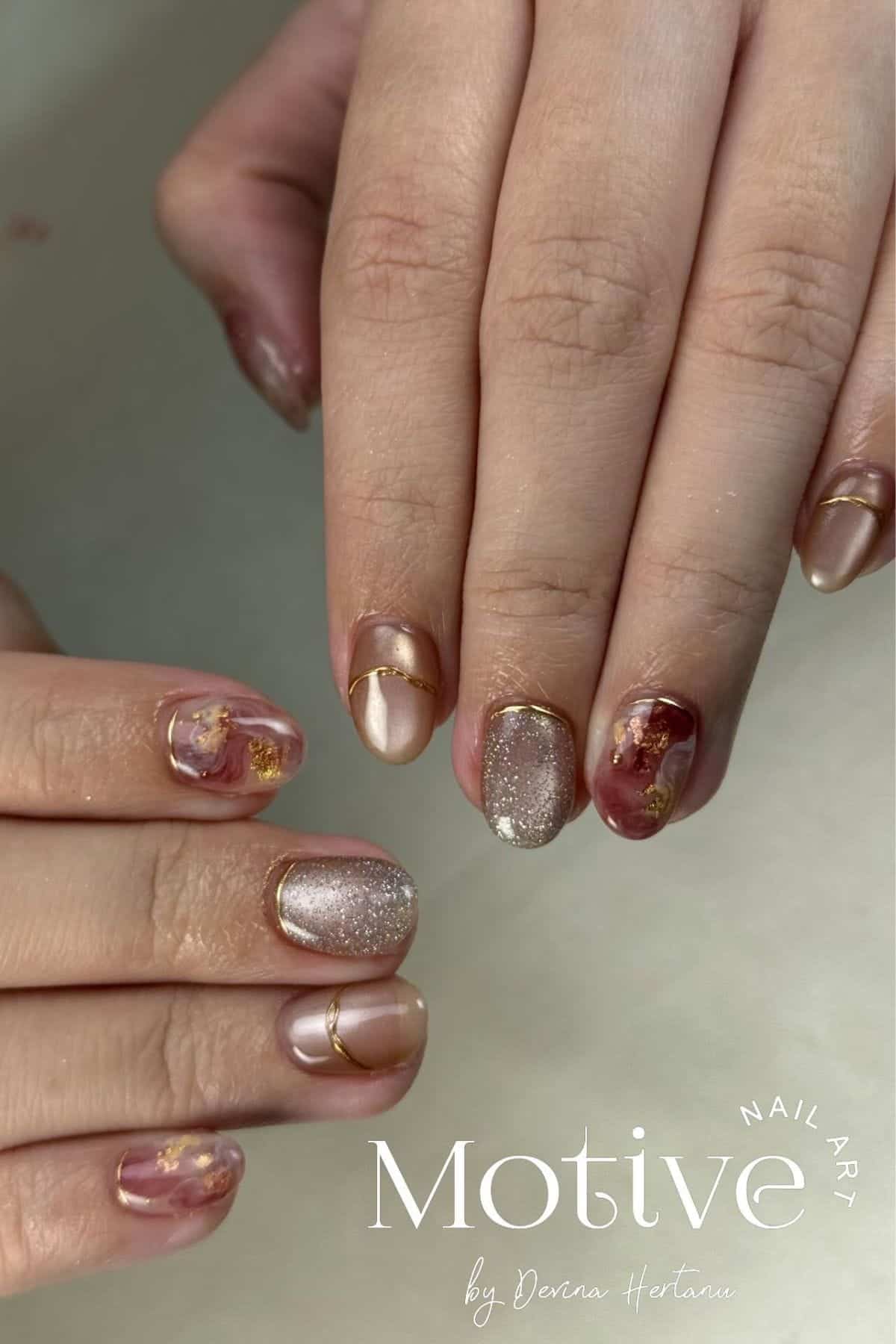 Close-up of hands with manicured nails featuring pink, gold, and glitter designs, including metallic accents and floral patterns. “Motive by Dena Hertanu” logo is visible in the corner.