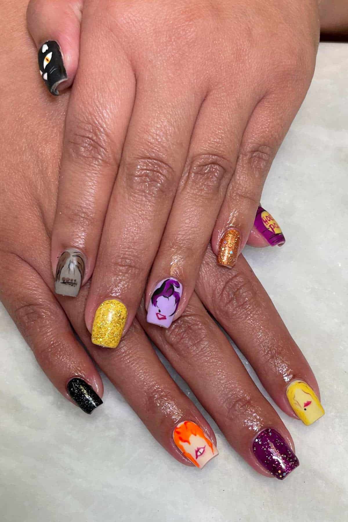 Hands with colorful, patterned nails featuring various illustrated faces and designs, including glitter and solid colors, resting on a white surface.