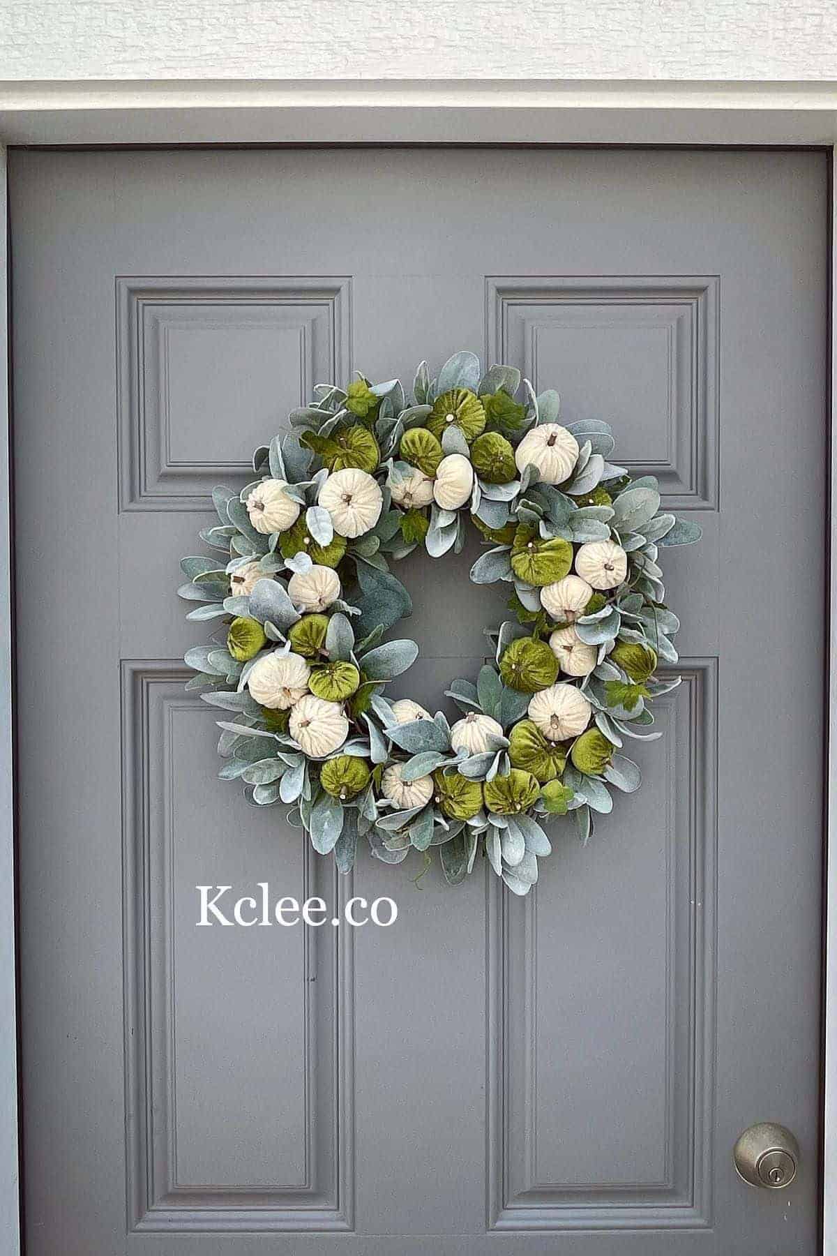 A wreath with green leaves and white pumpkins hangs on a gray door. The text “Kclee.co” is visible on the lower half of the wreath.