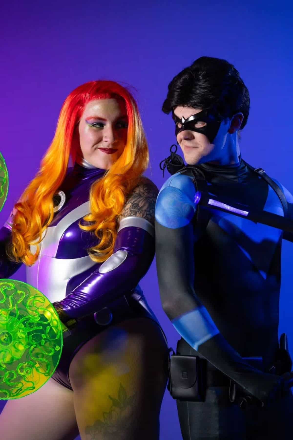 Two people in superhero costumes pose together; one wears a purple outfit with orange hair, the other wears a black suit with a mask and utility belt, both on a blue and purple gradient background.
