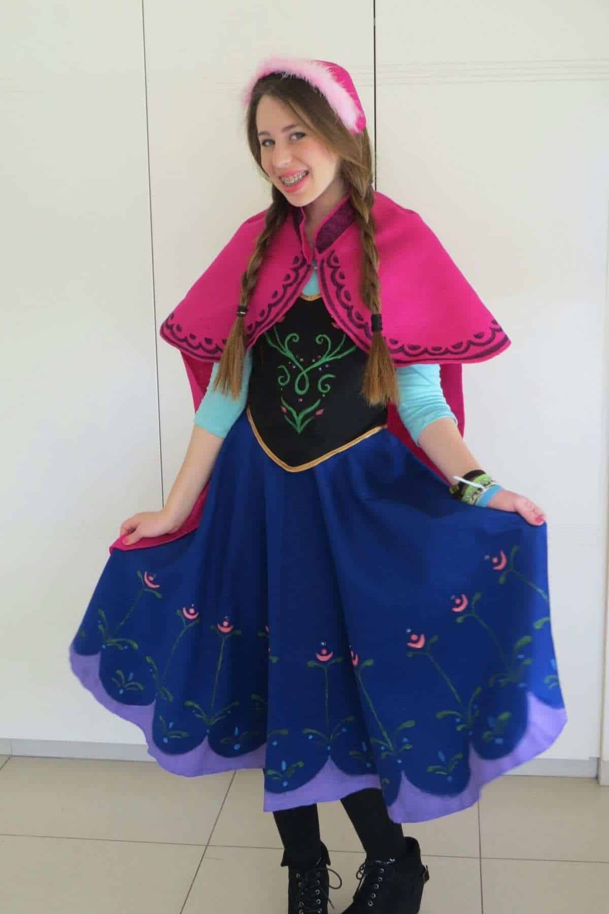 Person dressed in a blue and purple skirt, black vest, and pink cape, with braided hair and a pink headband, smiling and holding the sides of the skirt.