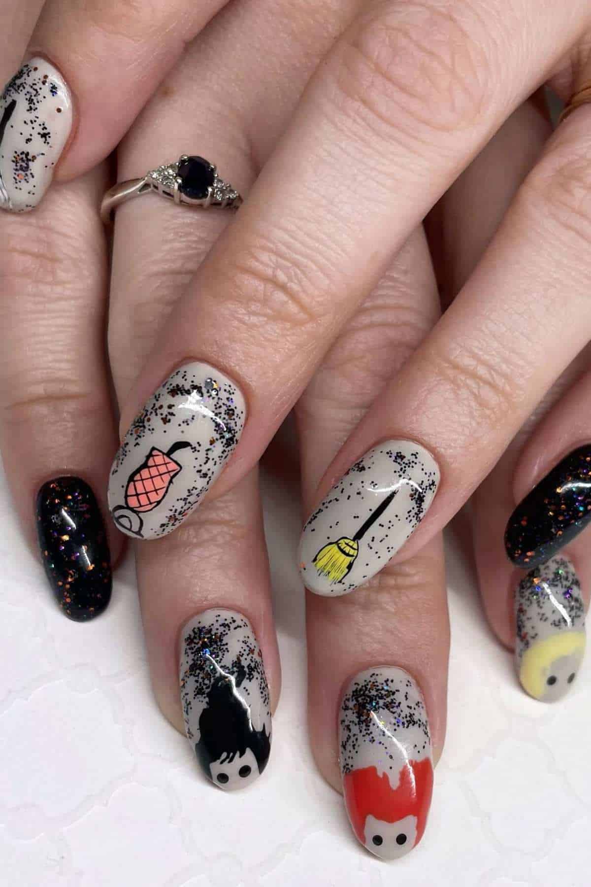 Hands with a ring and manicured nails featuring glitter and cartoon-like faces, a broom, a fishnet, and various colorful designs on a light background.