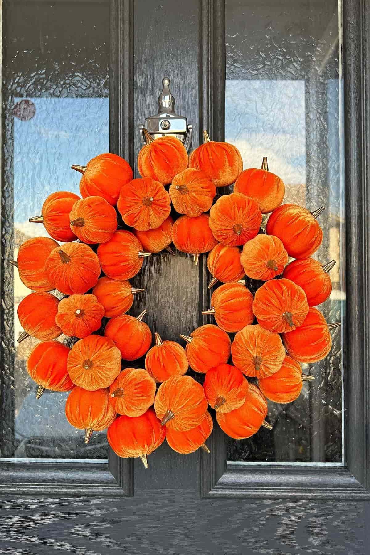 A round wreath made of small, bright orange pumpkins hangs on a dark front door with glass panels.