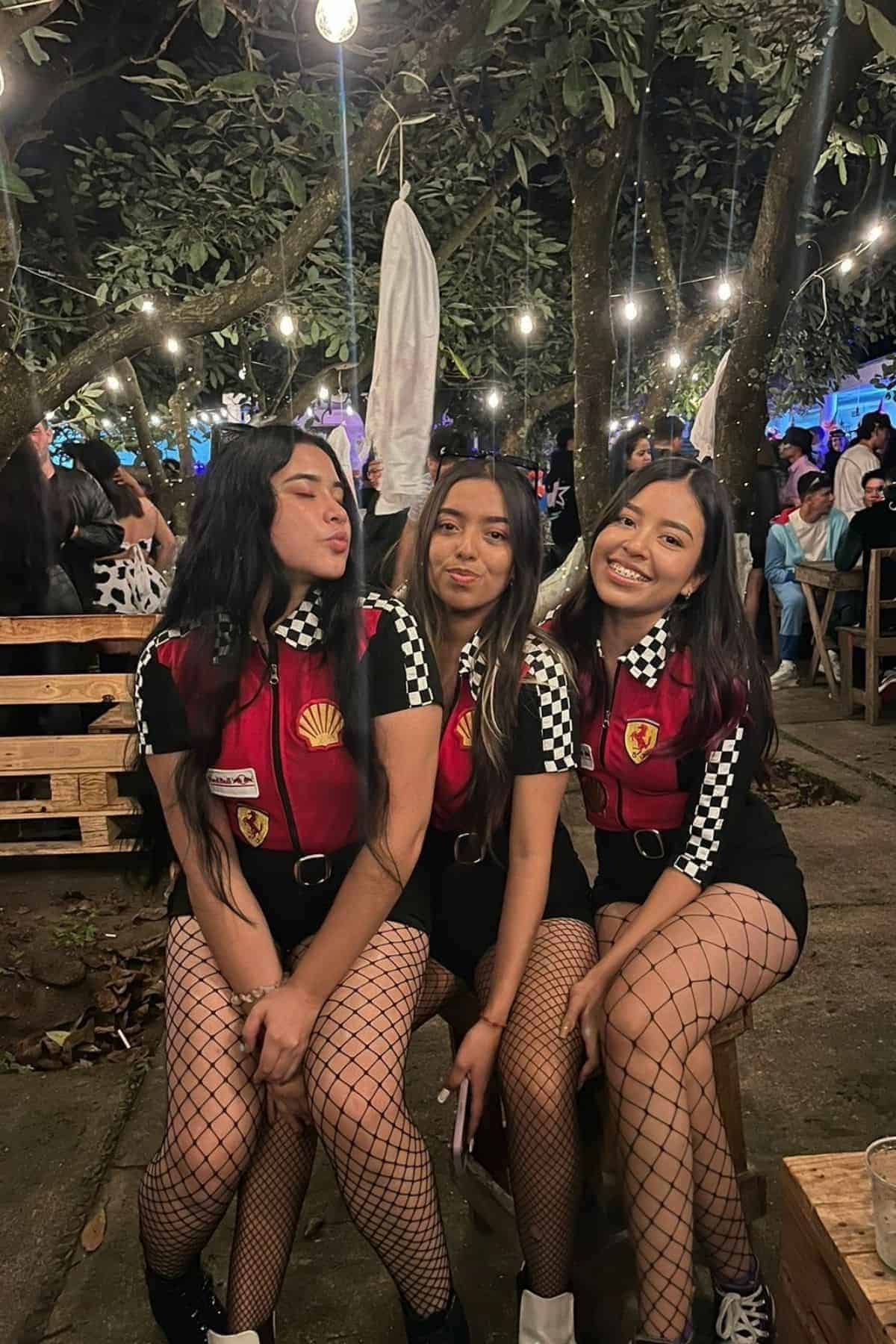 Three women in matching racing-inspired outfits and fishnet stockings sit on a bench outdoors at night, with string lights and other people in the background.