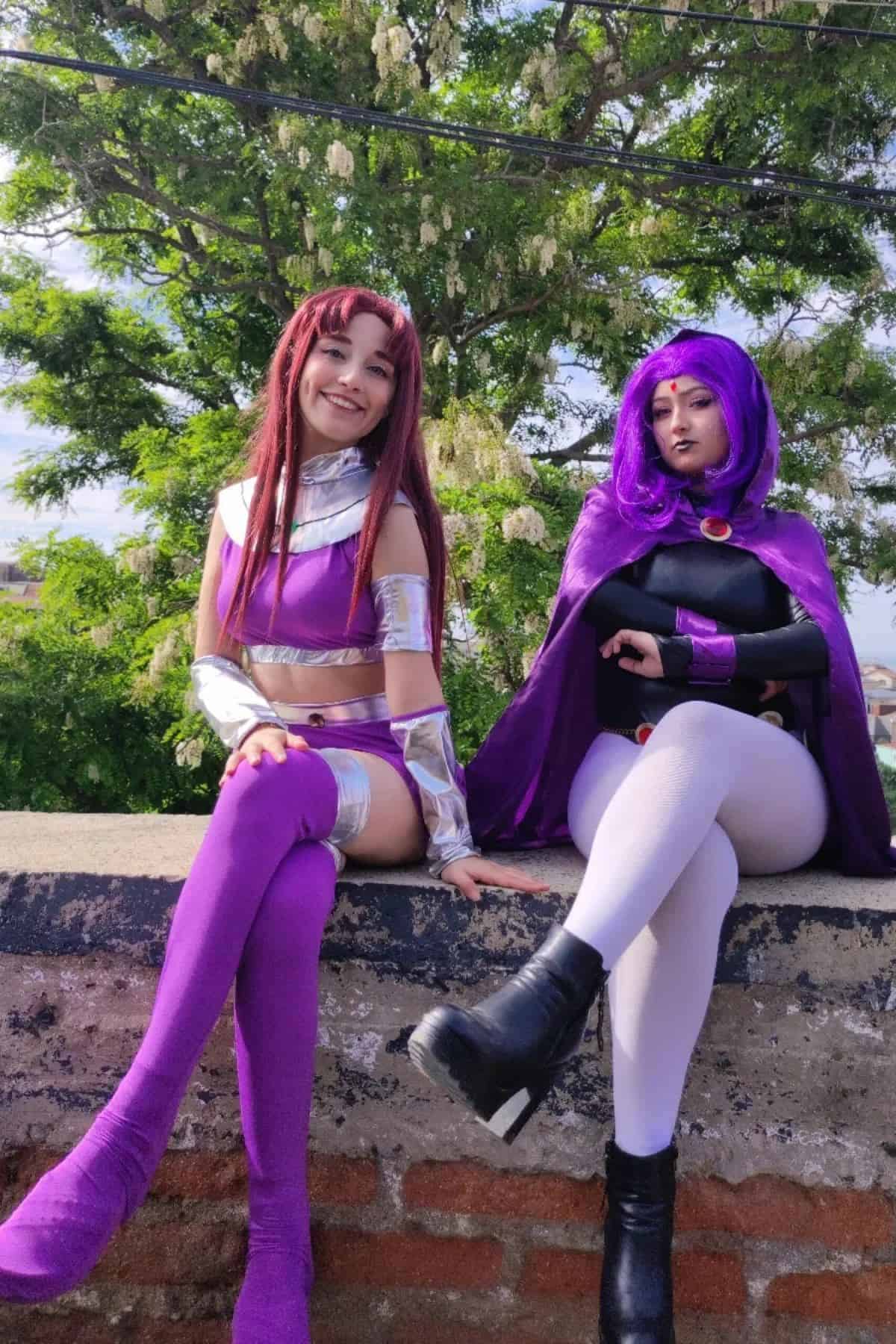 Two people in bright, detailed cosplay costumes pose sitting on a low brick wall outdoors, with green trees and sky in the background.