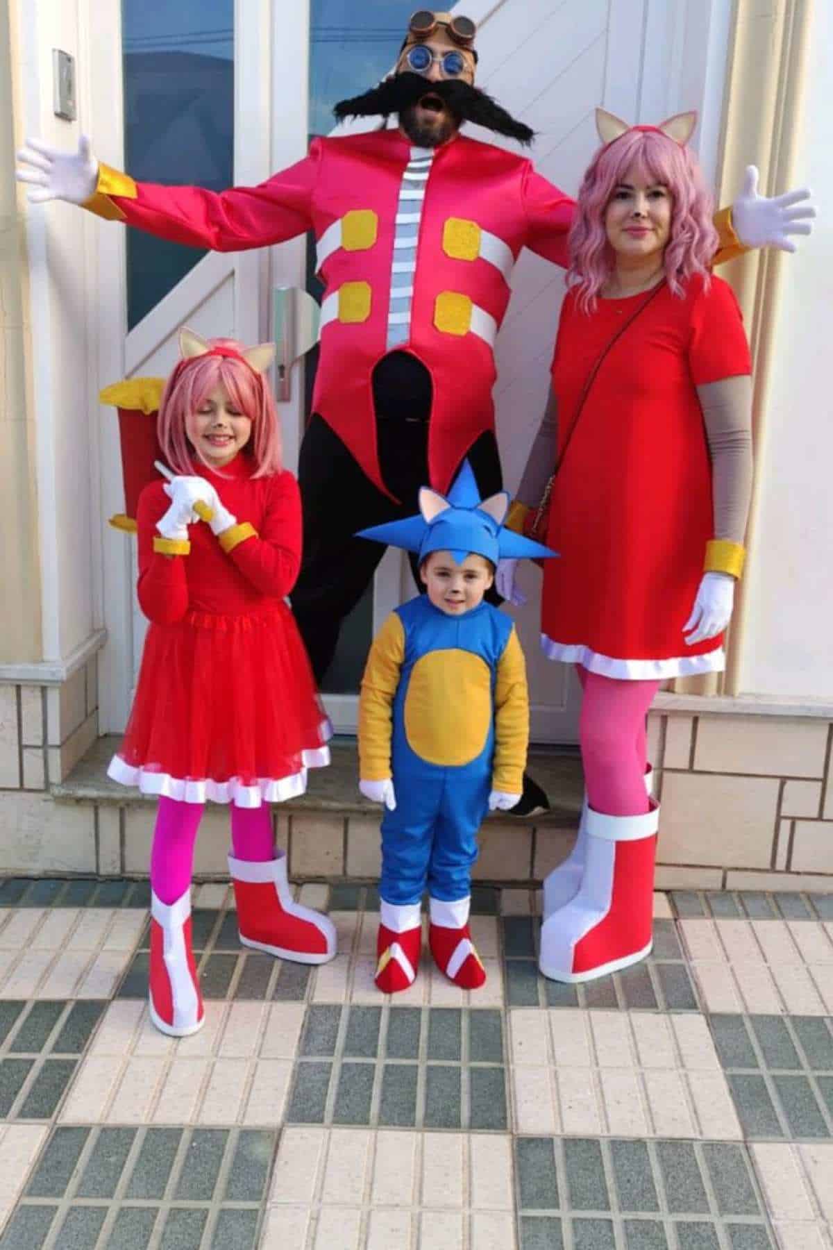 Four people dressed in Sonic the Hedgehog character costumes pose together outside on a tiled sidewalk in front of a white door.