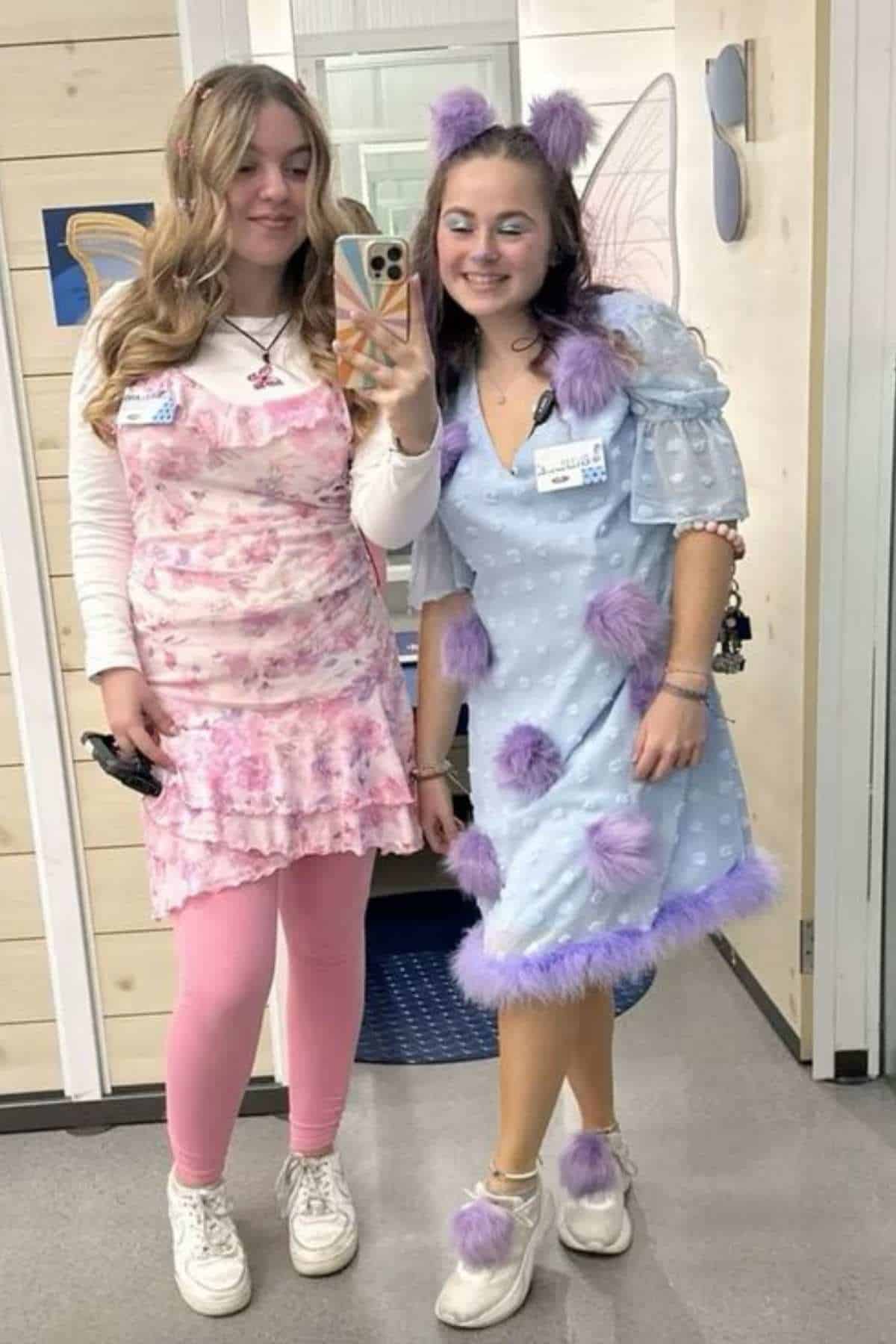 Two young women stand indoors, posing for a mirror selfie. One wears a pink floral outfit, the other a blue dress with purple fuzzy spots and matching headband. Both are smiling.