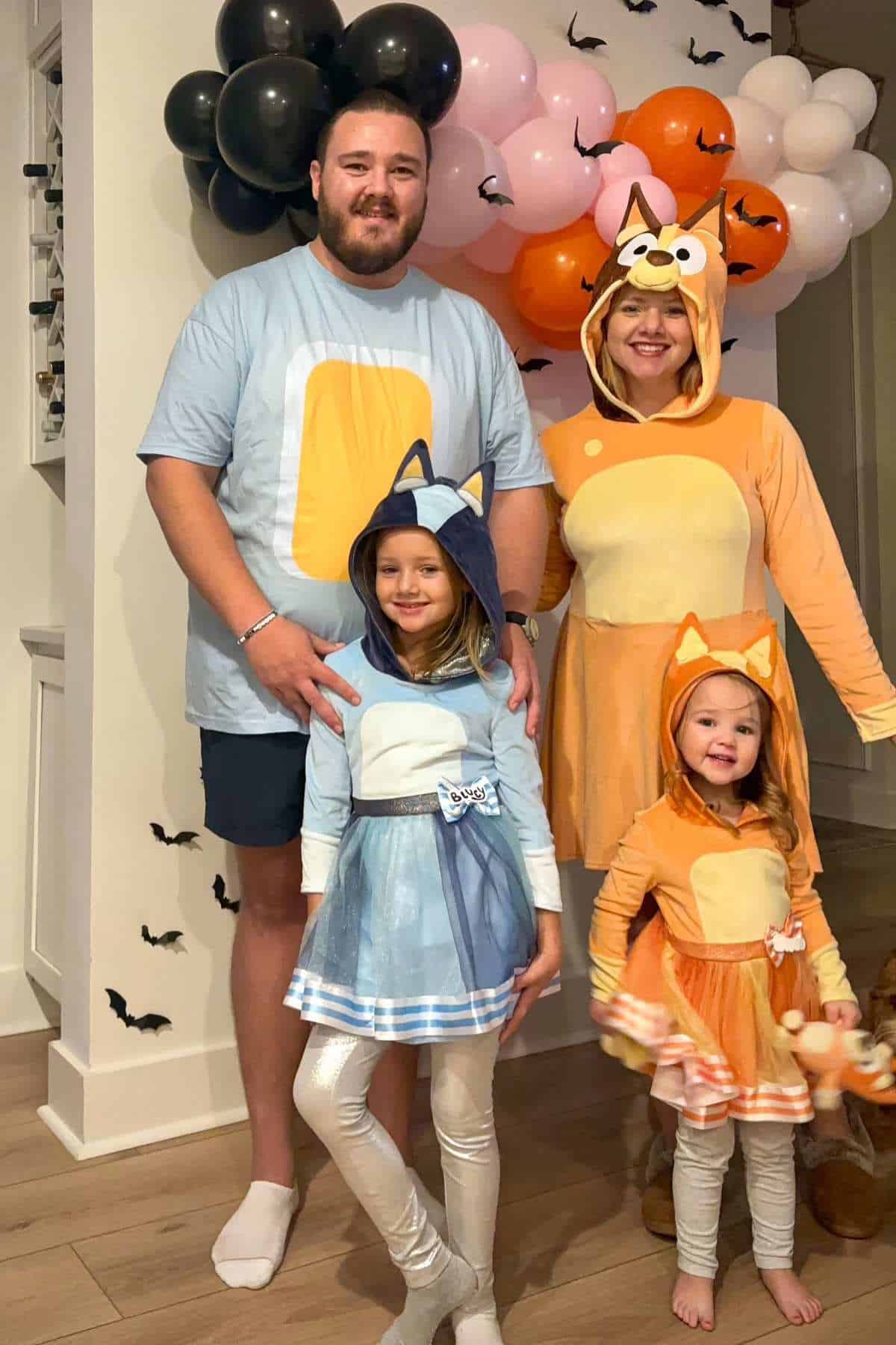 A family of four poses indoors in matching Bluey-themed costumes, with balloon decorations and black paper bats on the wall behind them.