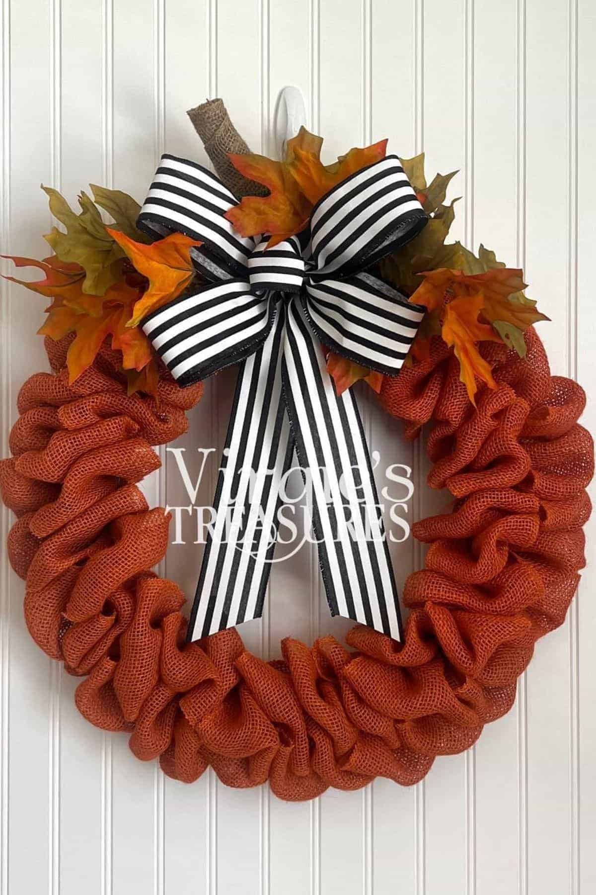 An orange burlap wreath with autumn leaves and a black-and-white striped bow hangs on a white paneled wall.
