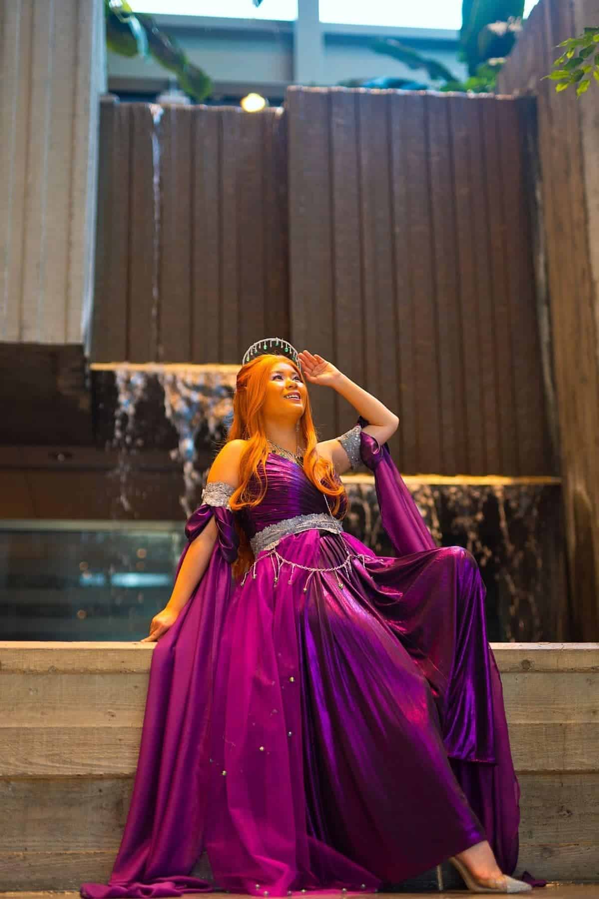 Person in a flowing purple dress and headpiece sits on stone steps in front of a cascading indoor waterfall, looking upward and smiling.