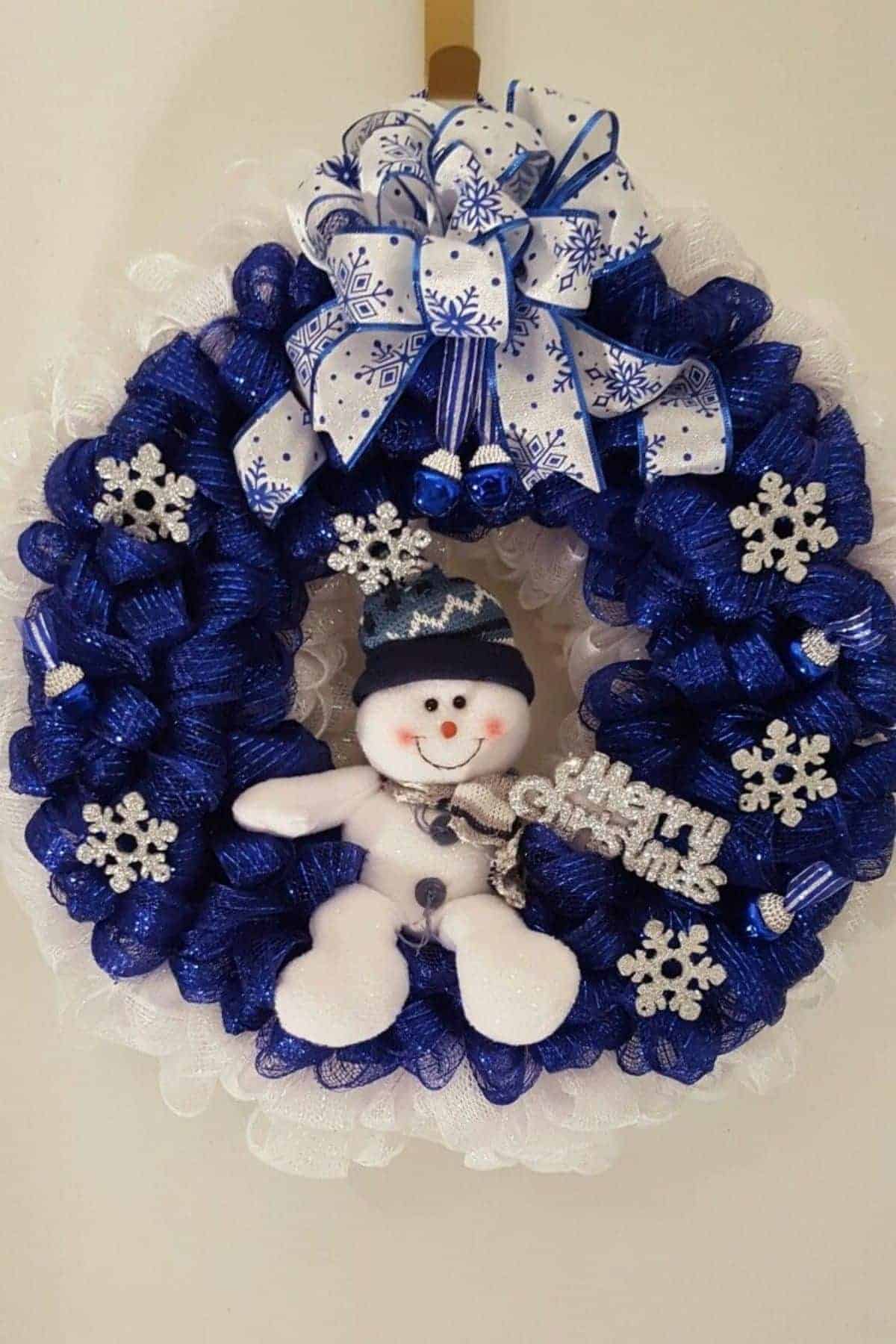 A blue and white wreath decorated with a snowman, snowflake ornaments, and a large bow with snowflake patterns.