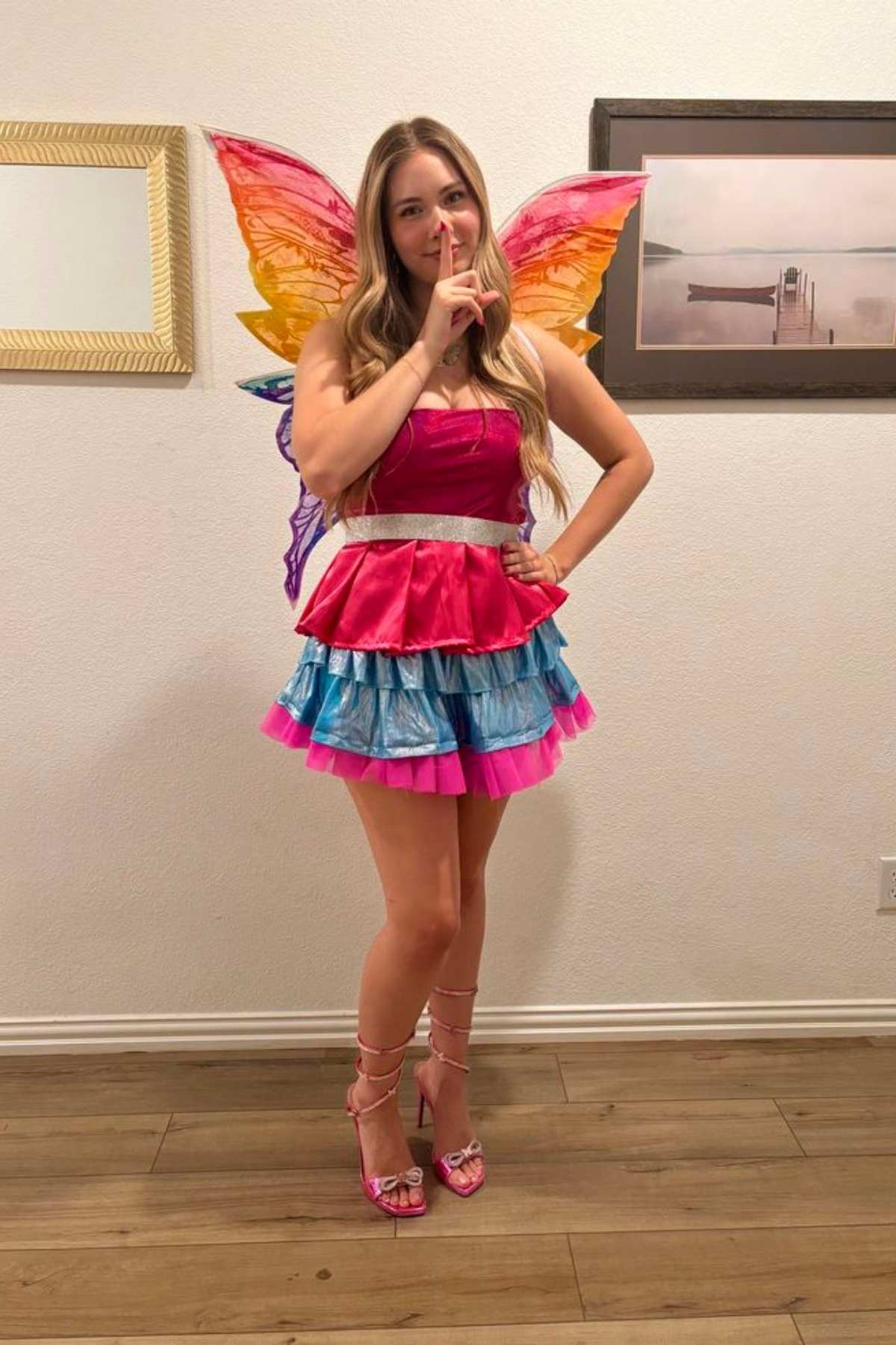 Woman wearing colorful fairy wings and a pink and blue tiered dress stands indoors, holding a finger to her lips in a “shh” gesture.