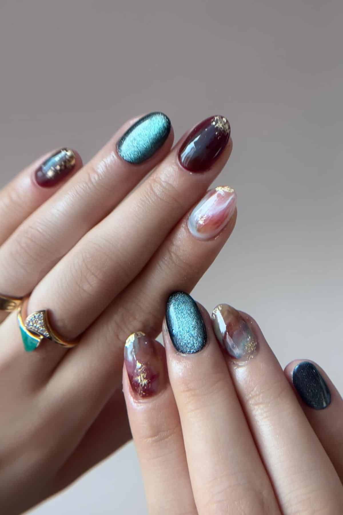 Hands with manicured nails in metallic teal, maroon, and marble designs with gold accents. Fingers are adorned with gold rings.