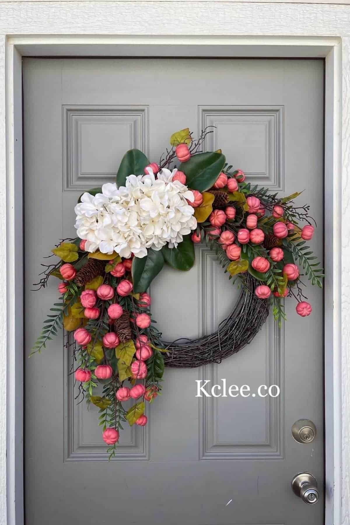 Wreath with white hydrangeas, pink buds, and green leaves on a gray door. The wreath is asymmetrical and the text “Kclee.co” appears in the lower right.