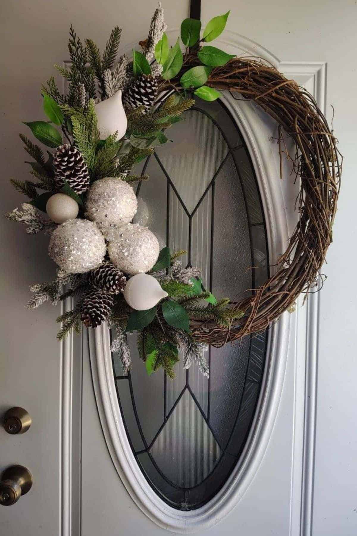 Half-decorated wreath with pinecones, white ornaments, artificial greenery, and branches hanging on an oval glass door.