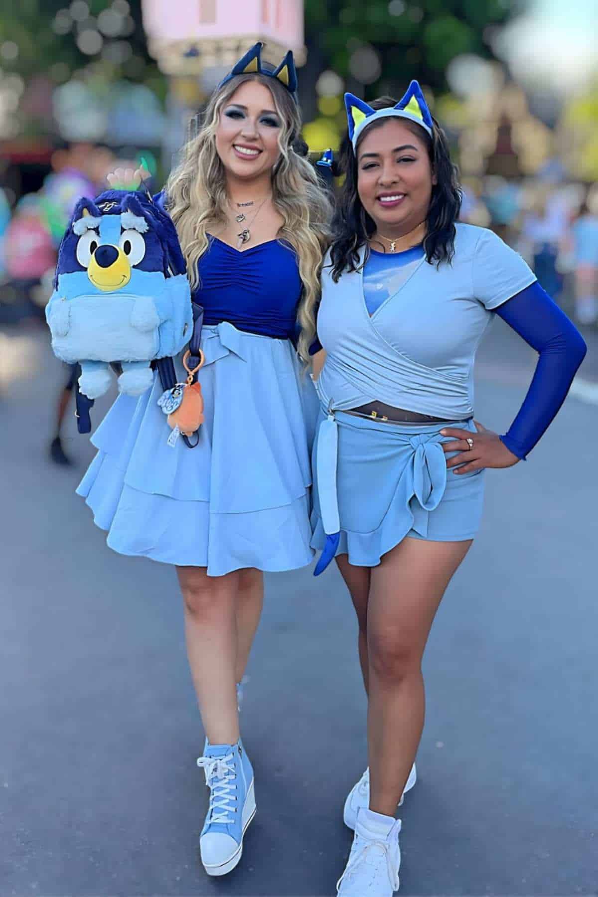 Two women wearing blue outfits and cat ear headbands pose together outdoors; one holds a matching blue character backpack.