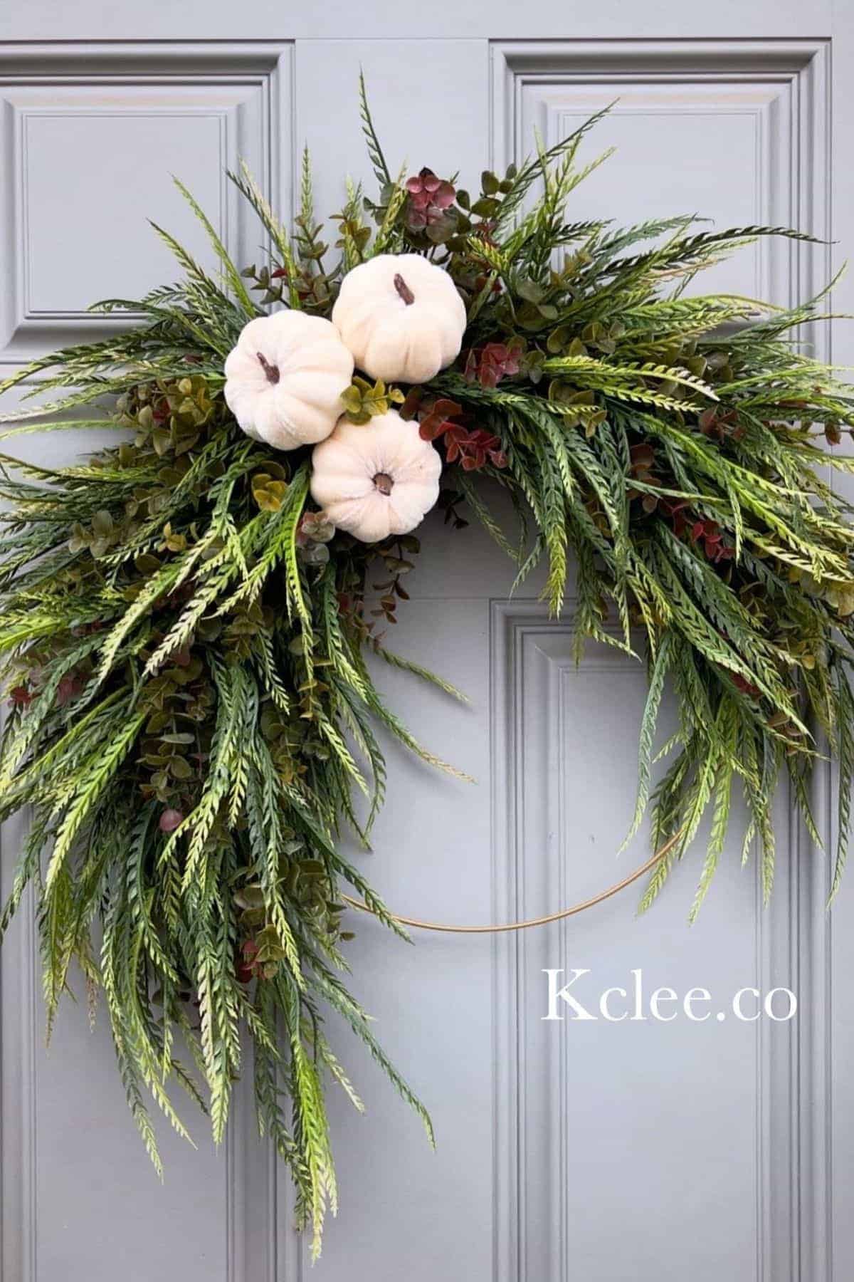 A wreath with green foliage and three white pumpkins hangs on a gray door.