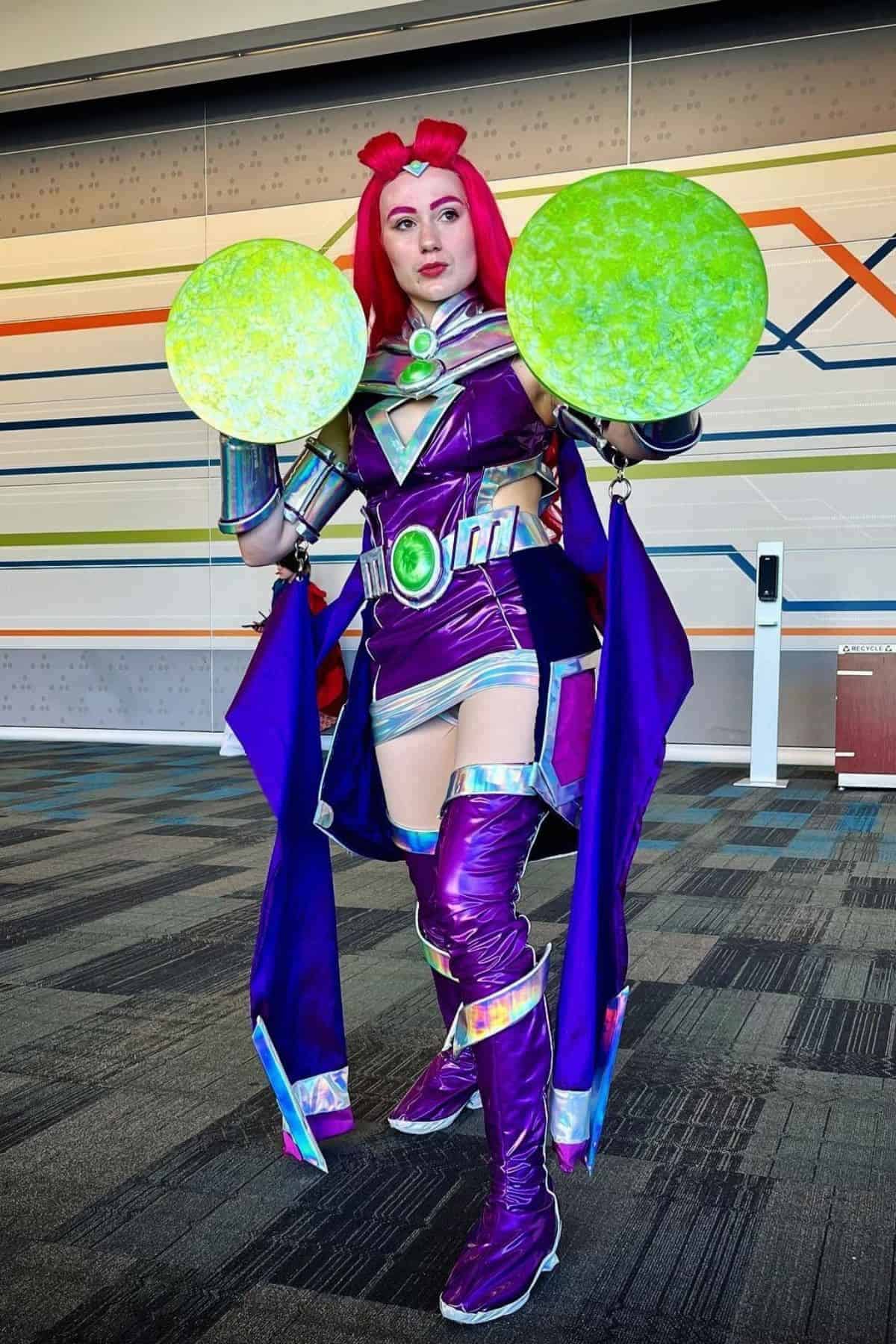 Person in a shiny purple and green costume with red hair, holding two large green discs, posing indoors against a wall with colorful lines.