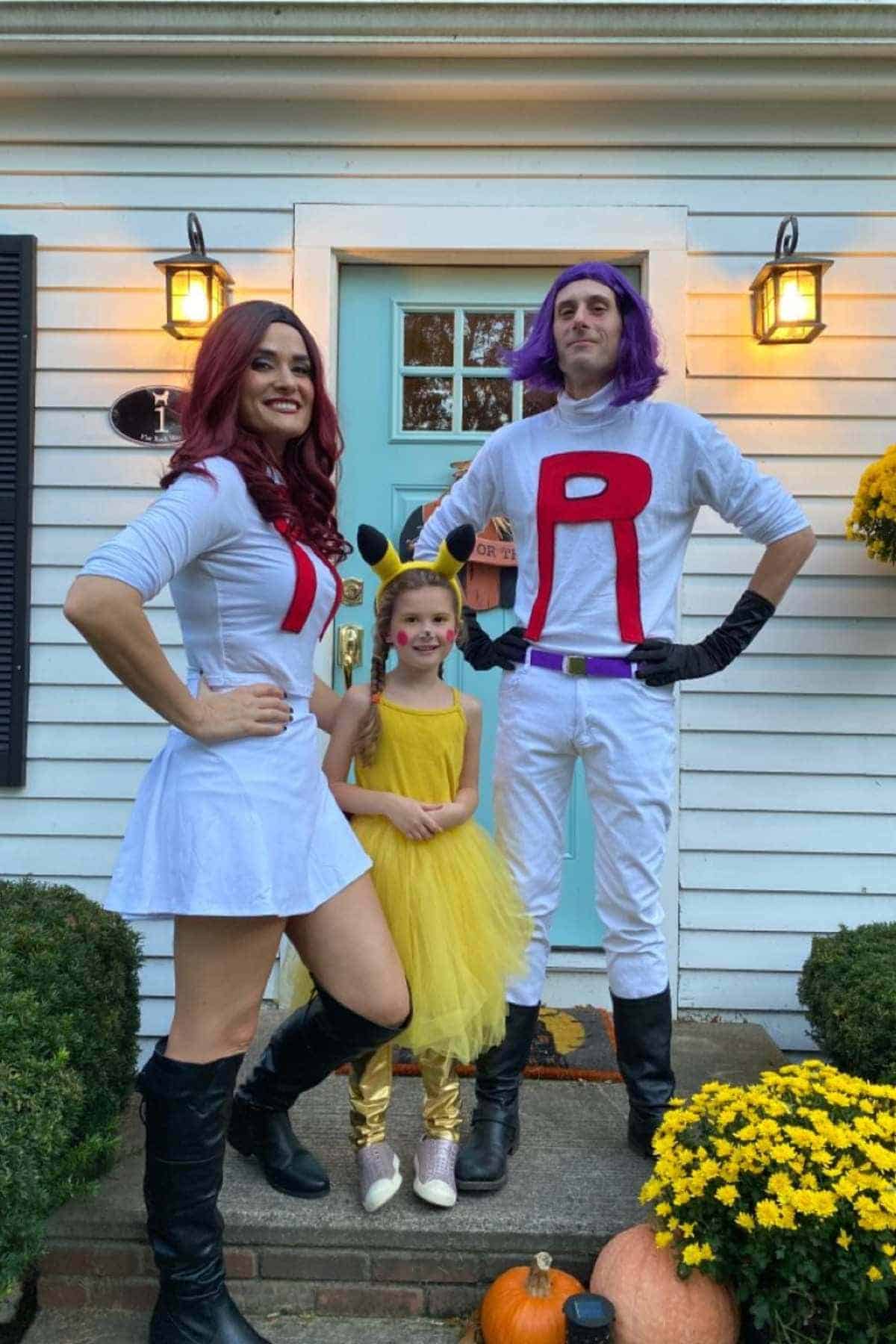 Two adults dressed as Team Rocket characters and a child dressed as Pikachu stand in front of a house decorated with pumpkins and yellow flowers.
