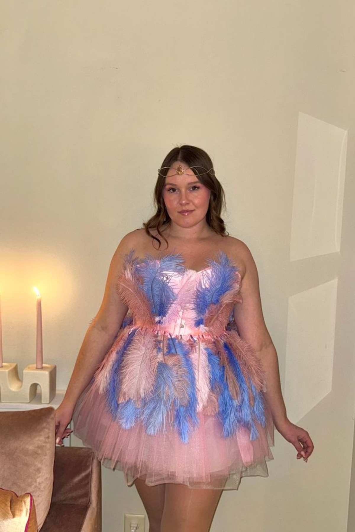 Woman stands indoors wearing a pink and blue feathered strapless dress, with a headpiece. Two lit candles on a table are visible in the background.