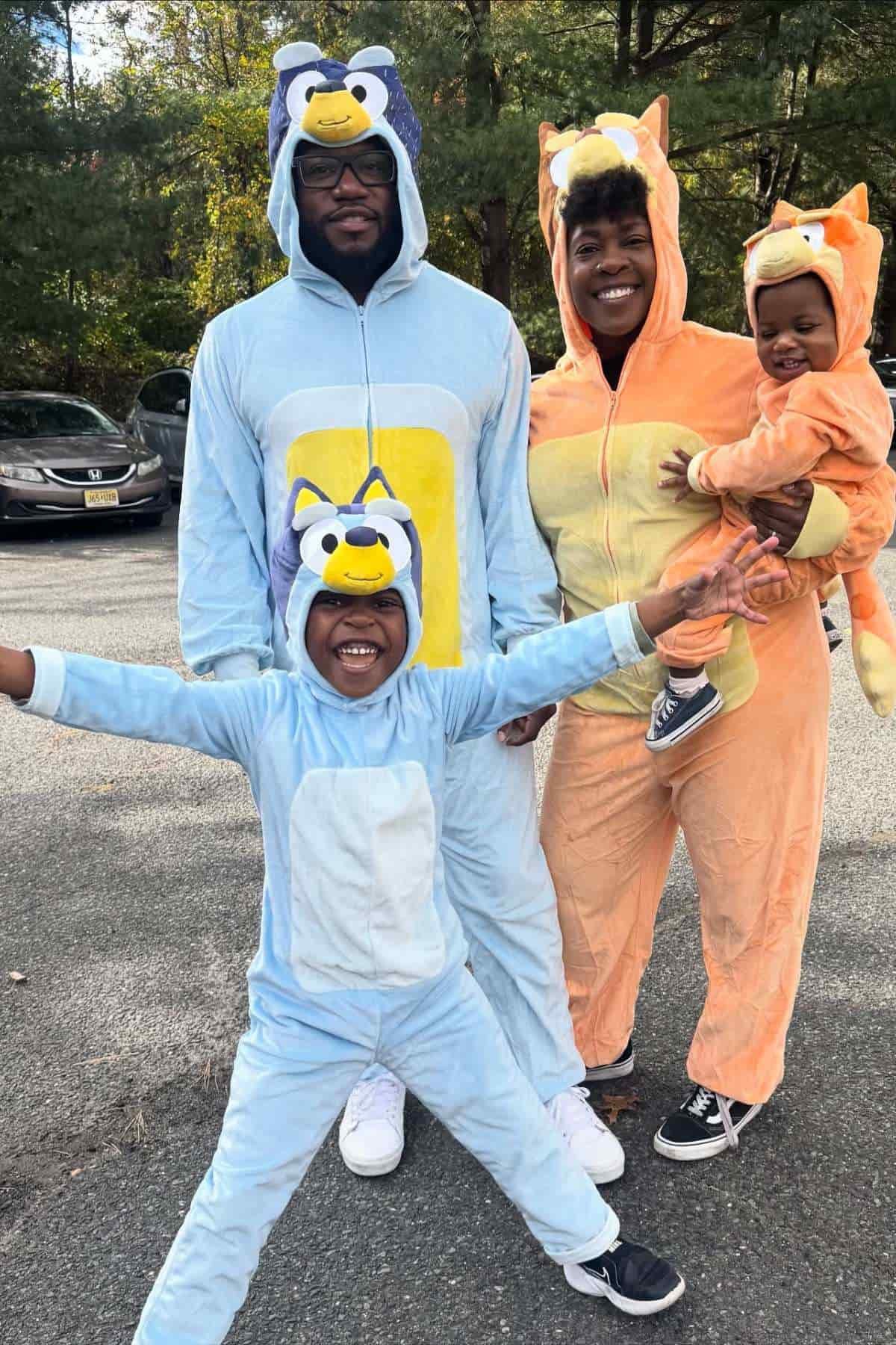 A family of four poses outdoors wearing colorful animal onesie costumes; two adults and two children, all smiling and happy.