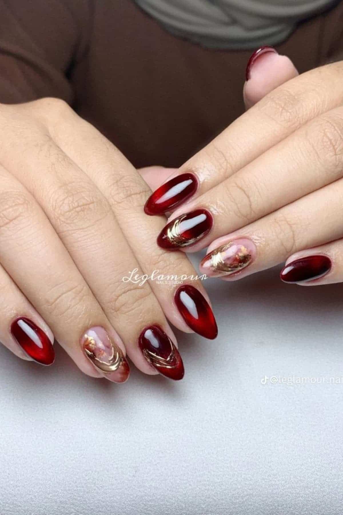 Hands with almond-shaped nails painted dark red, with accent nails featuring marble patterns and gold foil on a light background.