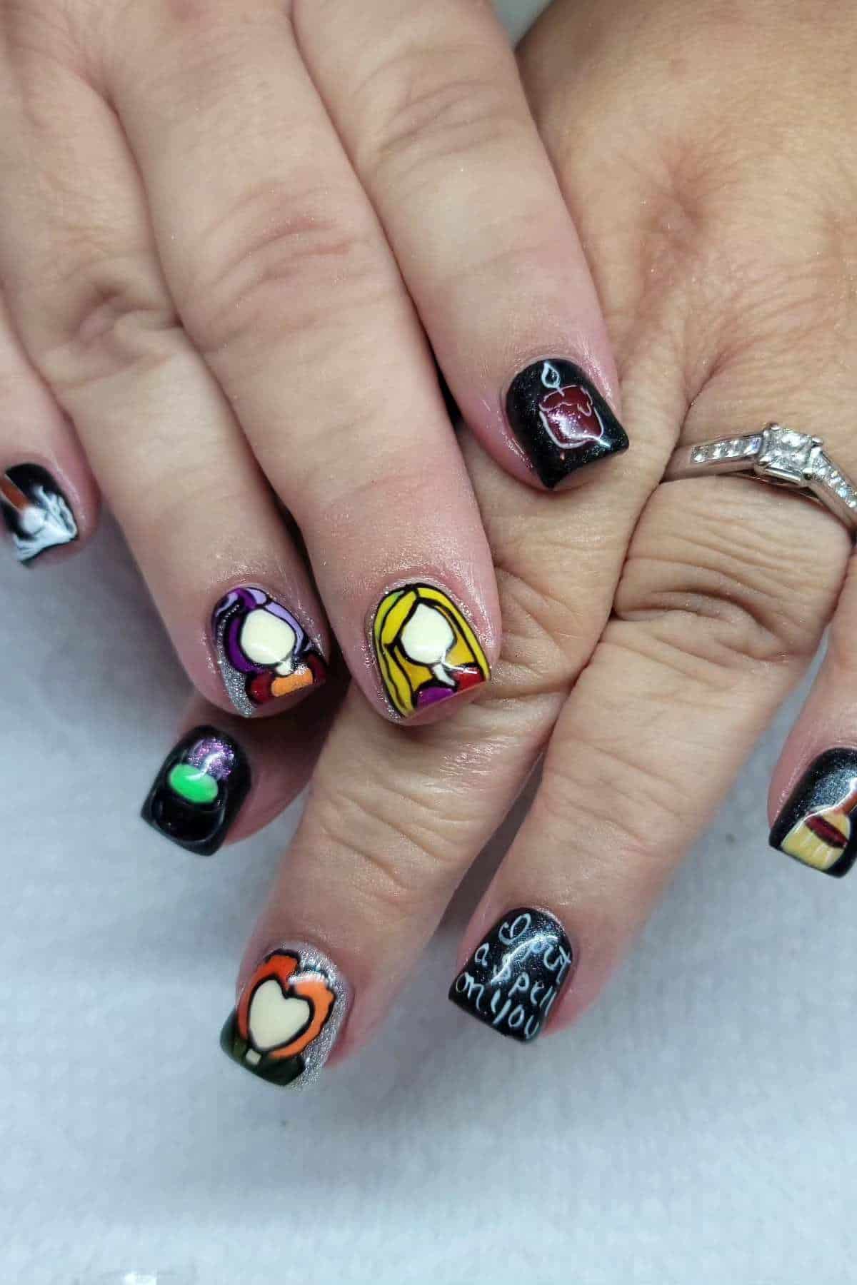 Hands with black nail polish featuring colorful pop art designs, including hearts, a woman’s face, lips, and the phrase “Got a spell on you” on one thumb.