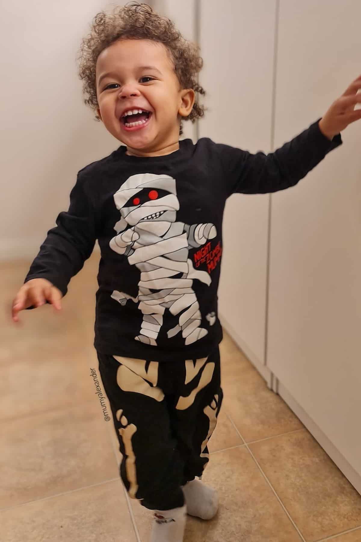 Smiling child wearing a black mummy-themed shirt and matching skeleton pants stands indoors on a tiled floor, with one arm raised and laughing.