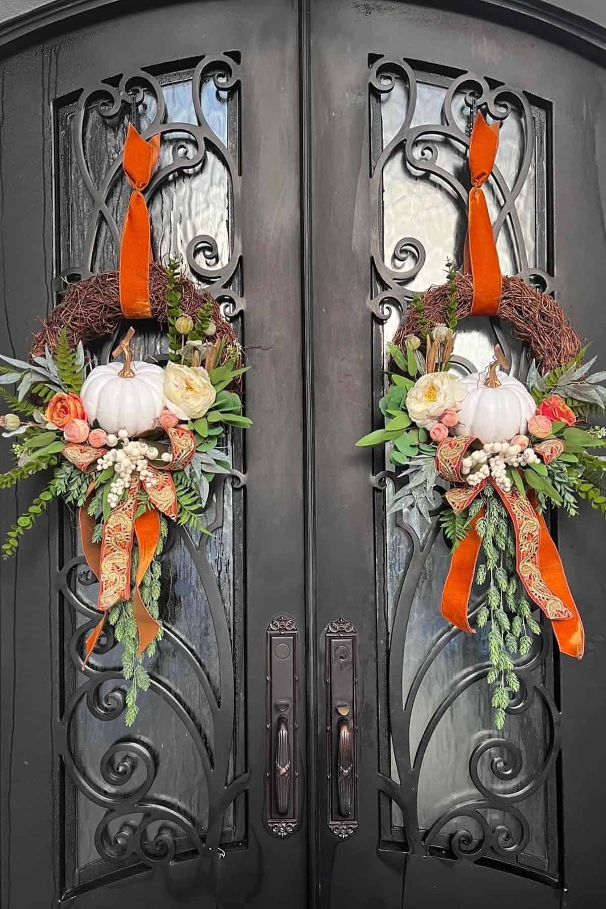 Two ornate dark double doors with identical autumn wreaths, featuring white pumpkins, orange ribbons, artificial flowers, and greenery.