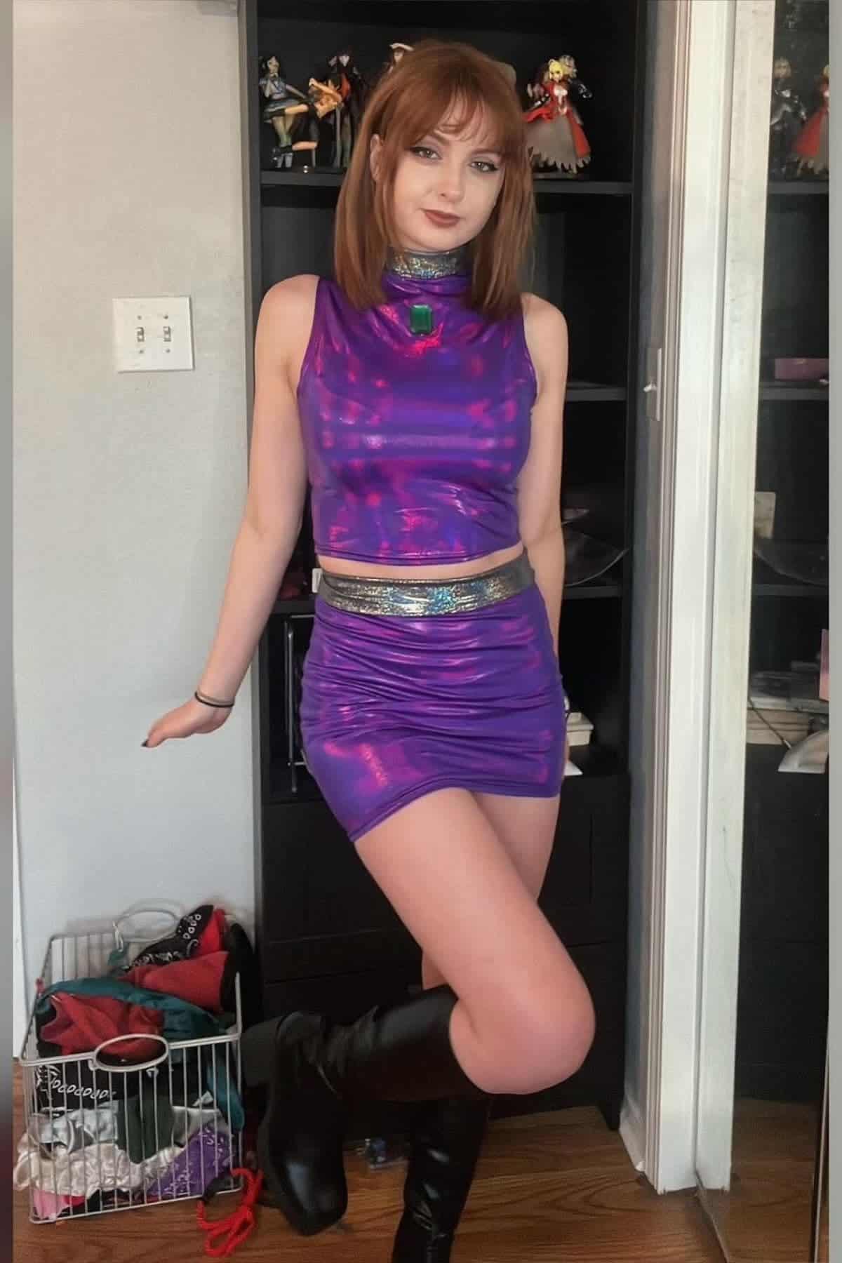 Woman standing indoors wearing a shiny purple sleeveless dress, black boots, and a choker, posing with one leg bent in front of a shelf with figures and an open basket on the floor.