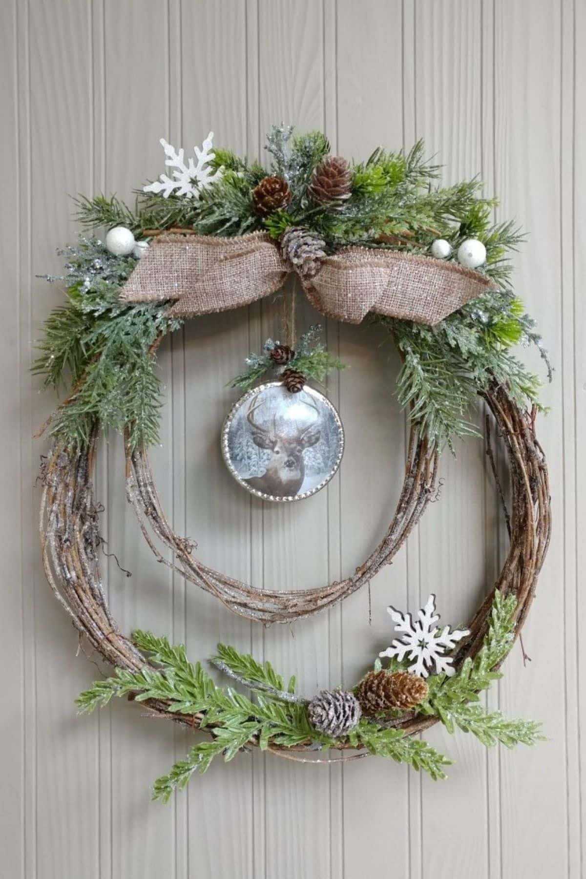 Double grapevine wreath decorated with pinecones, faux greenery, burlap bow, white ornaments, snowflakes, and a glass bauble, hanging on a light wooden wall.