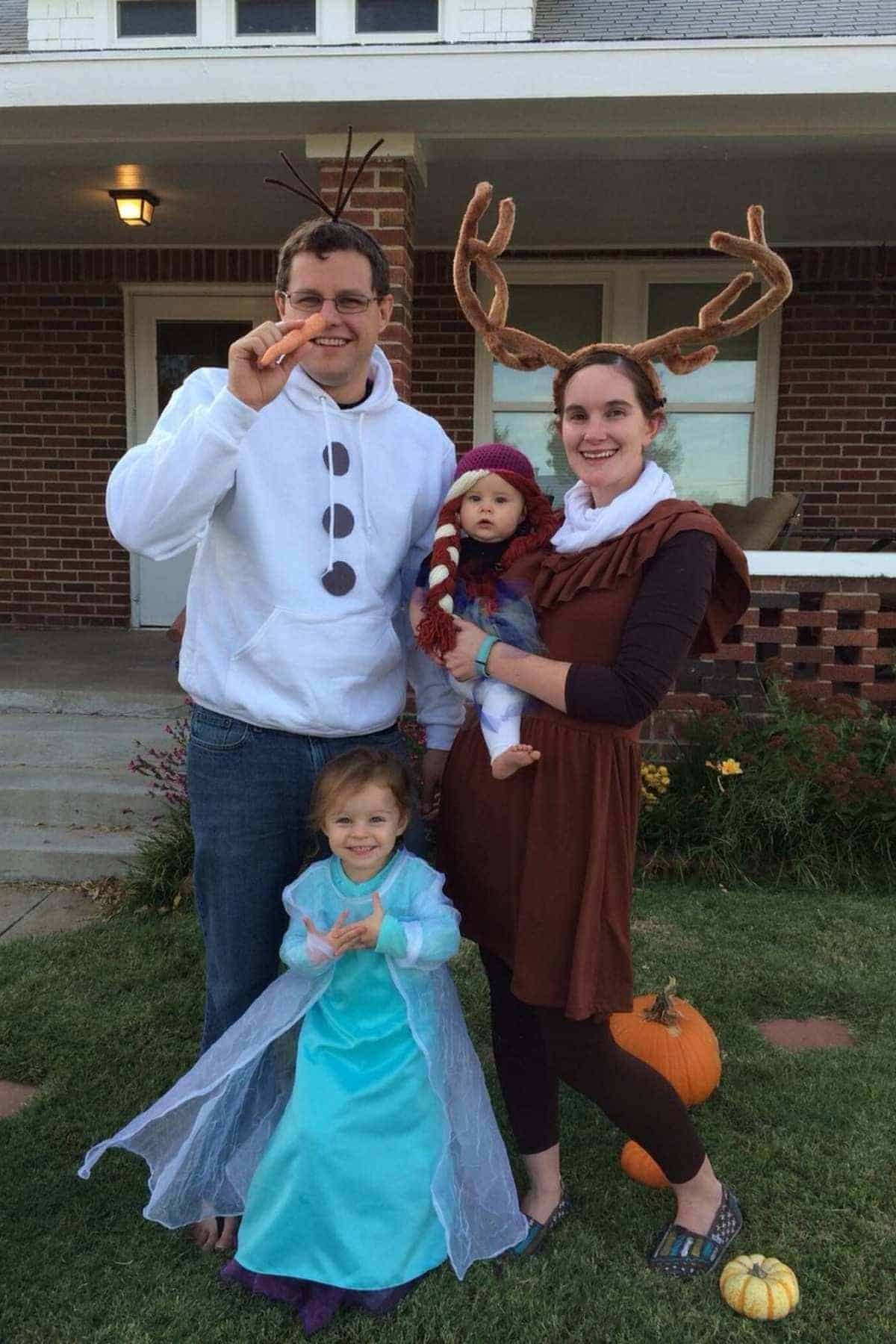 A family of four in “Frozen” costumes stands outside a house. The adults wear reindeer and snowman outfits, while the children are dressed as Elsa and another character.