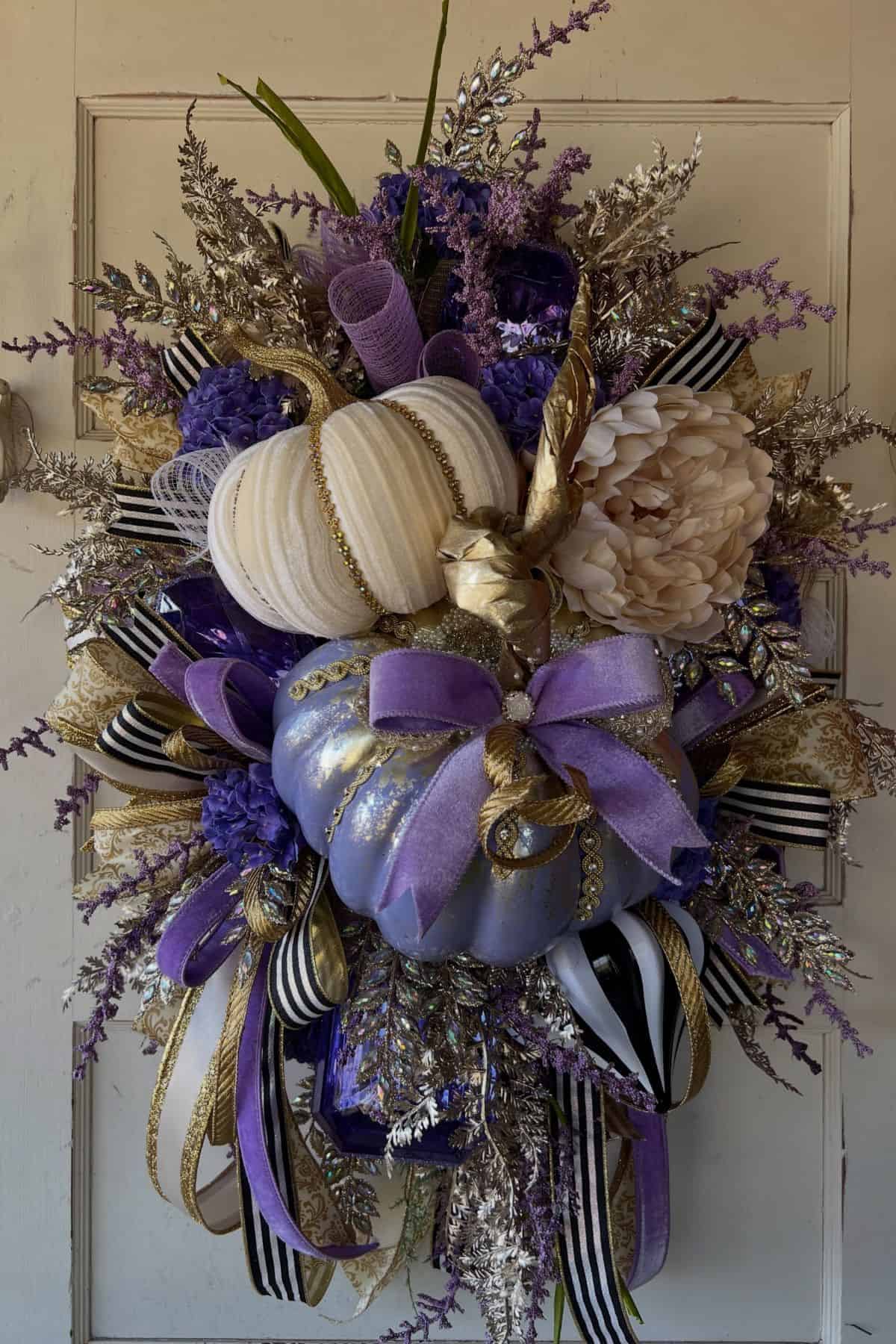 Decorative wreath featuring white and purple pumpkins, gold and purple bows, cream flower, green leaves, metallic and striped ribbons, and purple, gold, and beige foliage on a white door.