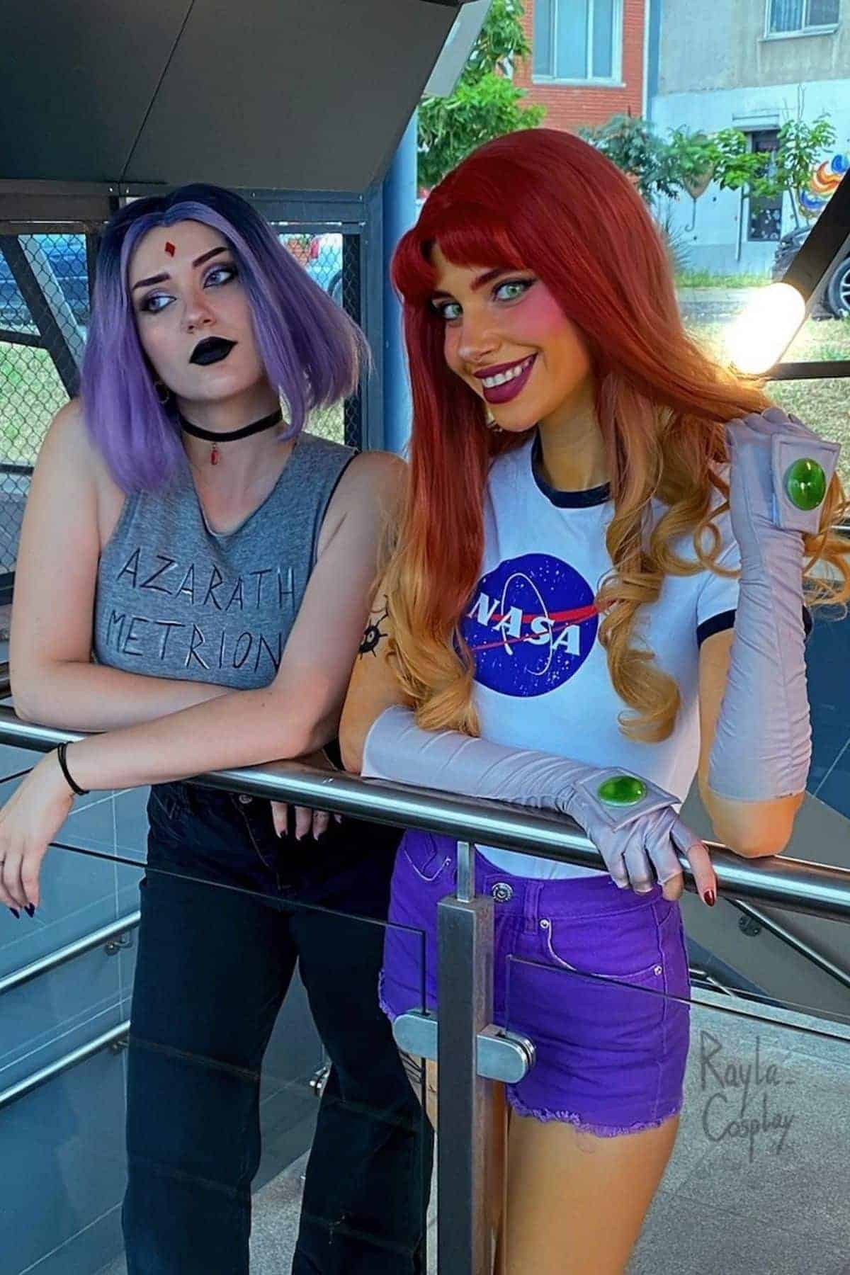 Two women pose in cosplay; one with short purple hair and a gray “Azarath Metrion” shirt, the other with long red hair, a NASA shirt, and purple shorts, both standing by a metal railing.